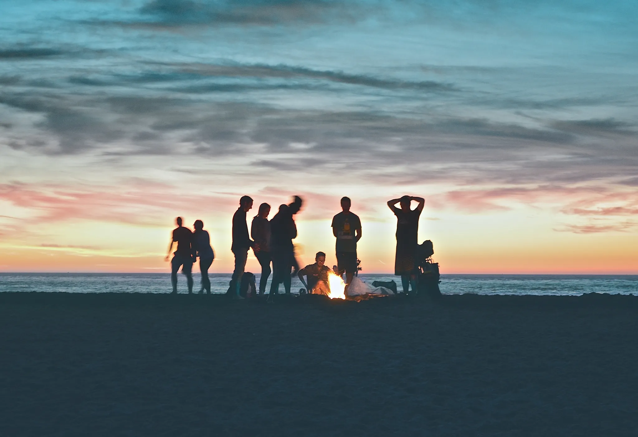 Silhouetted group of friends gathered around a beach bonfire at sunset with the ocean in the background.