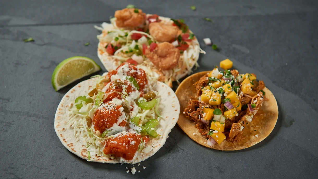 Three Rubio's tacos featuring tempura shrimp, salsa-topped fish, and mango-pork on a slate surface with lime wedge.