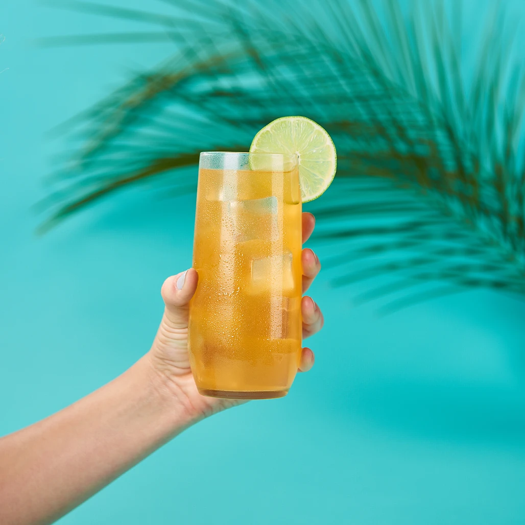 Hand holding a tall glass of iced tea with lime garnish in front of a palm frond on a turquoise backdrop.