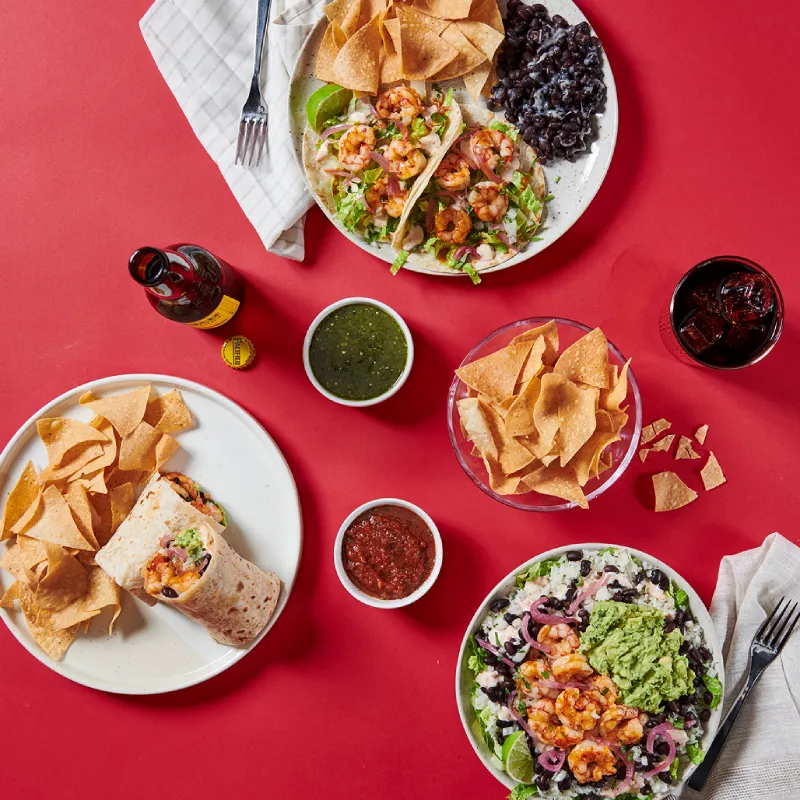 Overhead spread of Rubio's shrimp tacos, burrito with chips, salsas, and a shrimp bowl on a red table.