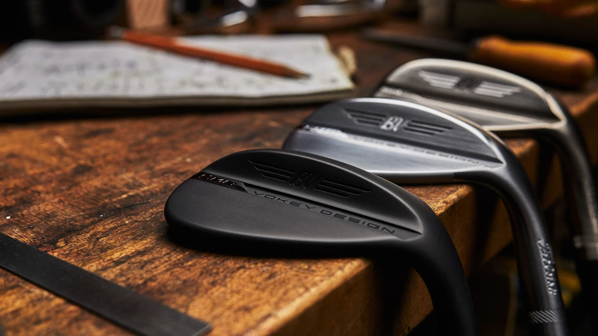 Three Vokey SM8 wedges in black, brushed steel, and chrome finishes lined up on a workbench beside design sketches.