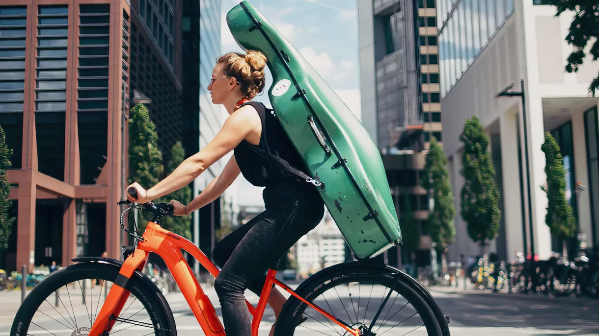 Cyclist in city traffic riding an orange Cannondale bike with a green cello case strapped to her back past modern office buildings.