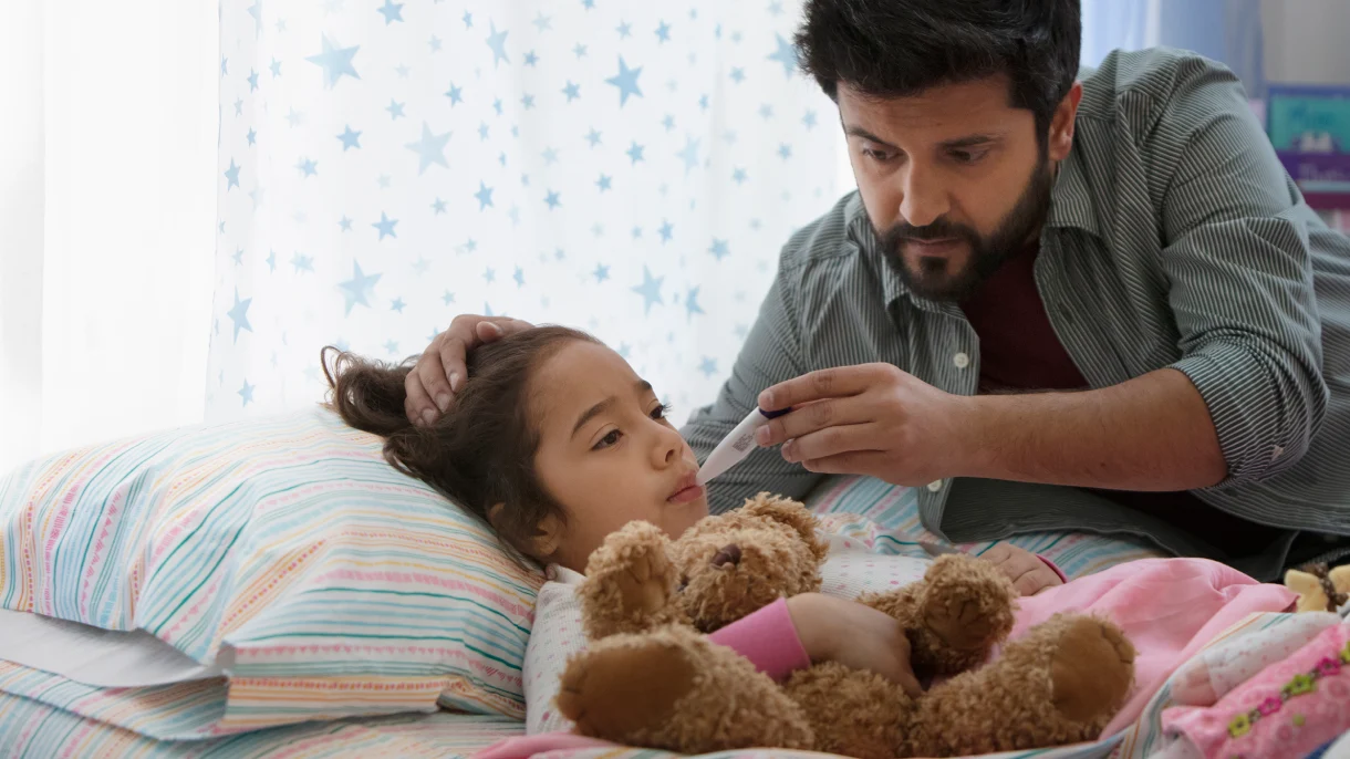 A caring man checks the temperature of a sick child lying in bed, who is holding a teddy bear. The scene conveys concern and tenderness.