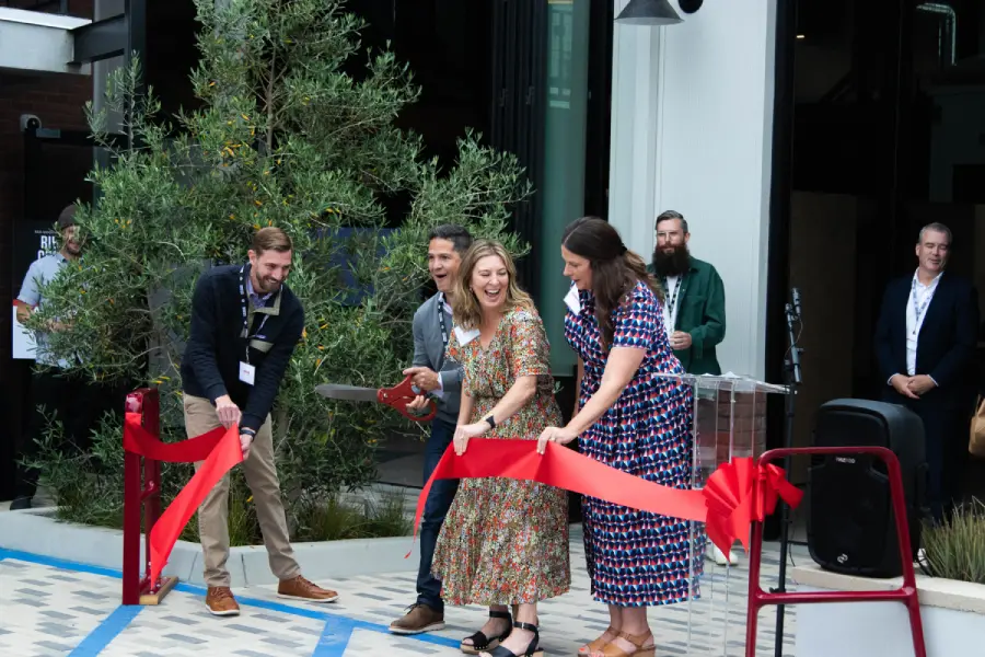 Three smiling people cut a red ribbon with oversized scissors at a grand opening ceremony outside a modern building, as colleagues look on.