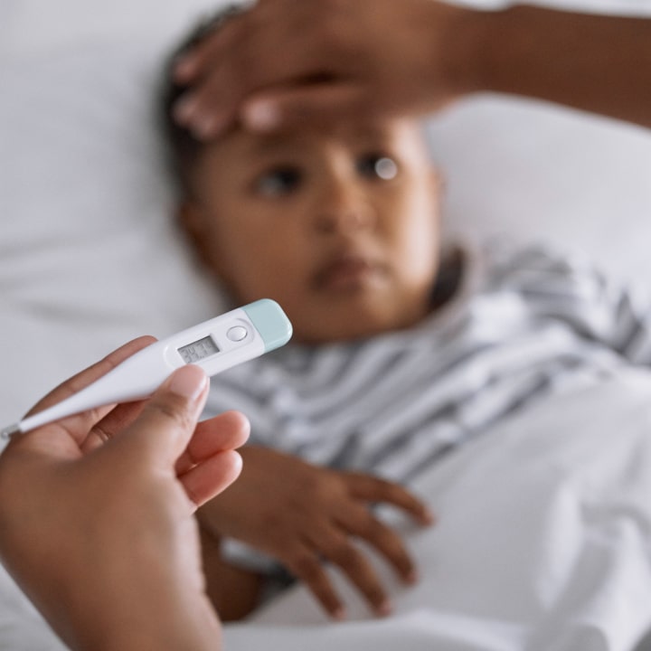A worried child lies in bed with a hand on their forehead, while someone holds a digital thermometer in the foreground, indicating a fever check.