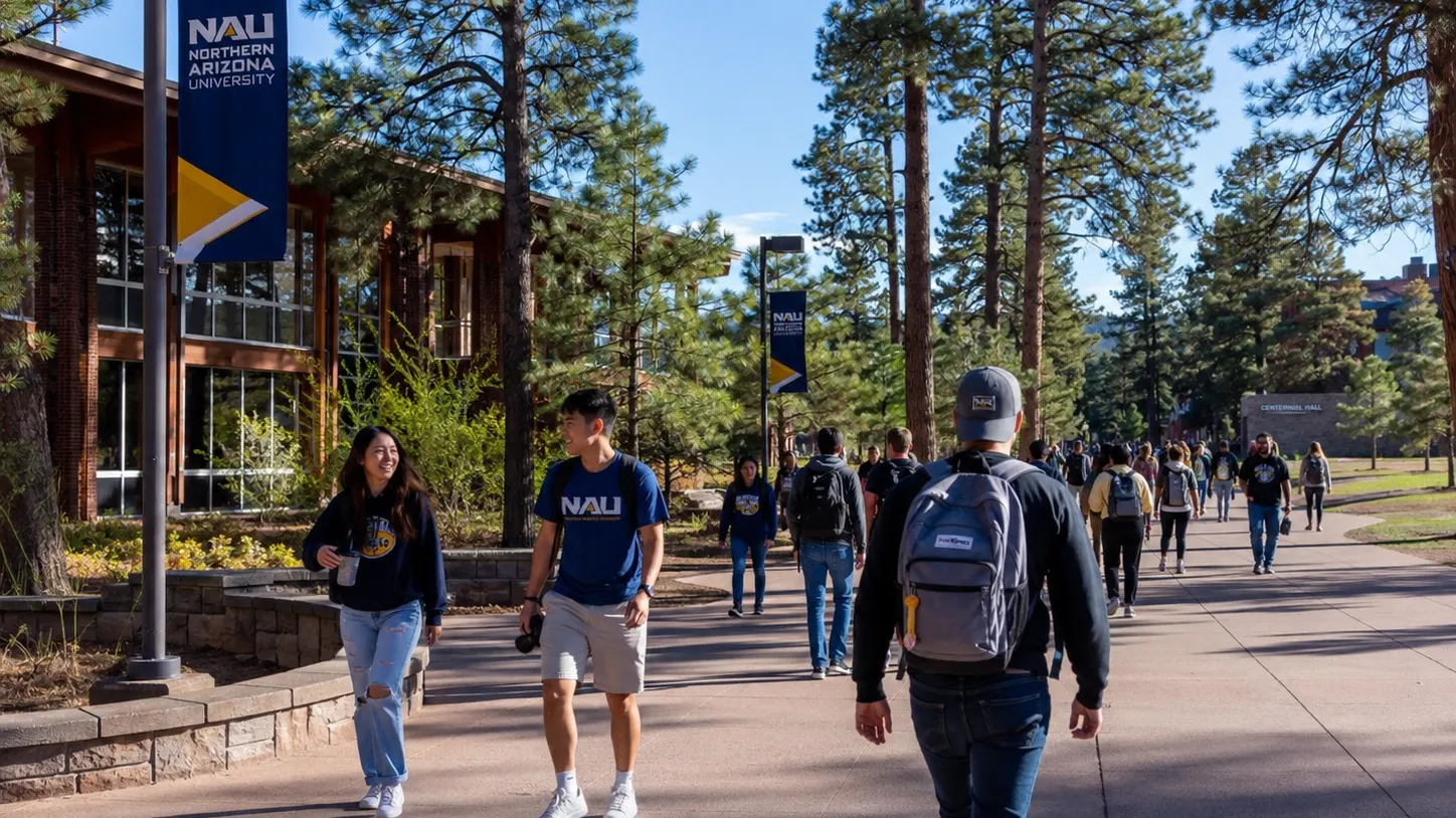 Students walk along a pine-lined Northern Arizona University campus pathway on a bright, sunny day.