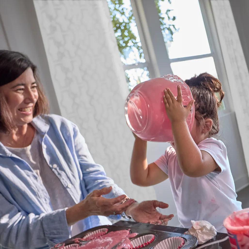 A woman laughs as a child licks pink batter from a bowl, with cupcake trays in front, sunlight streaming through windows.