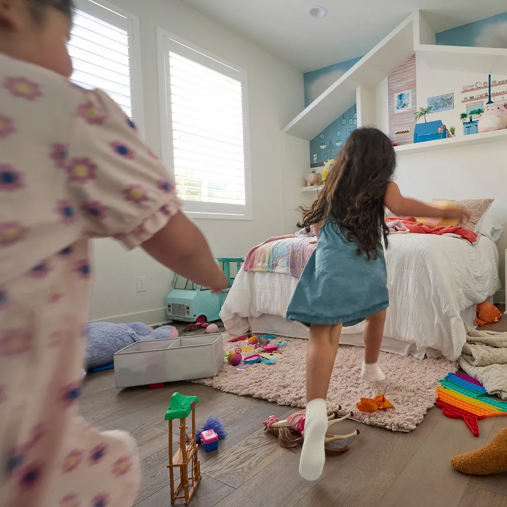 Two children play energetically in a colorful, toy-filled bedroom with a cozy bed and creative wall decor