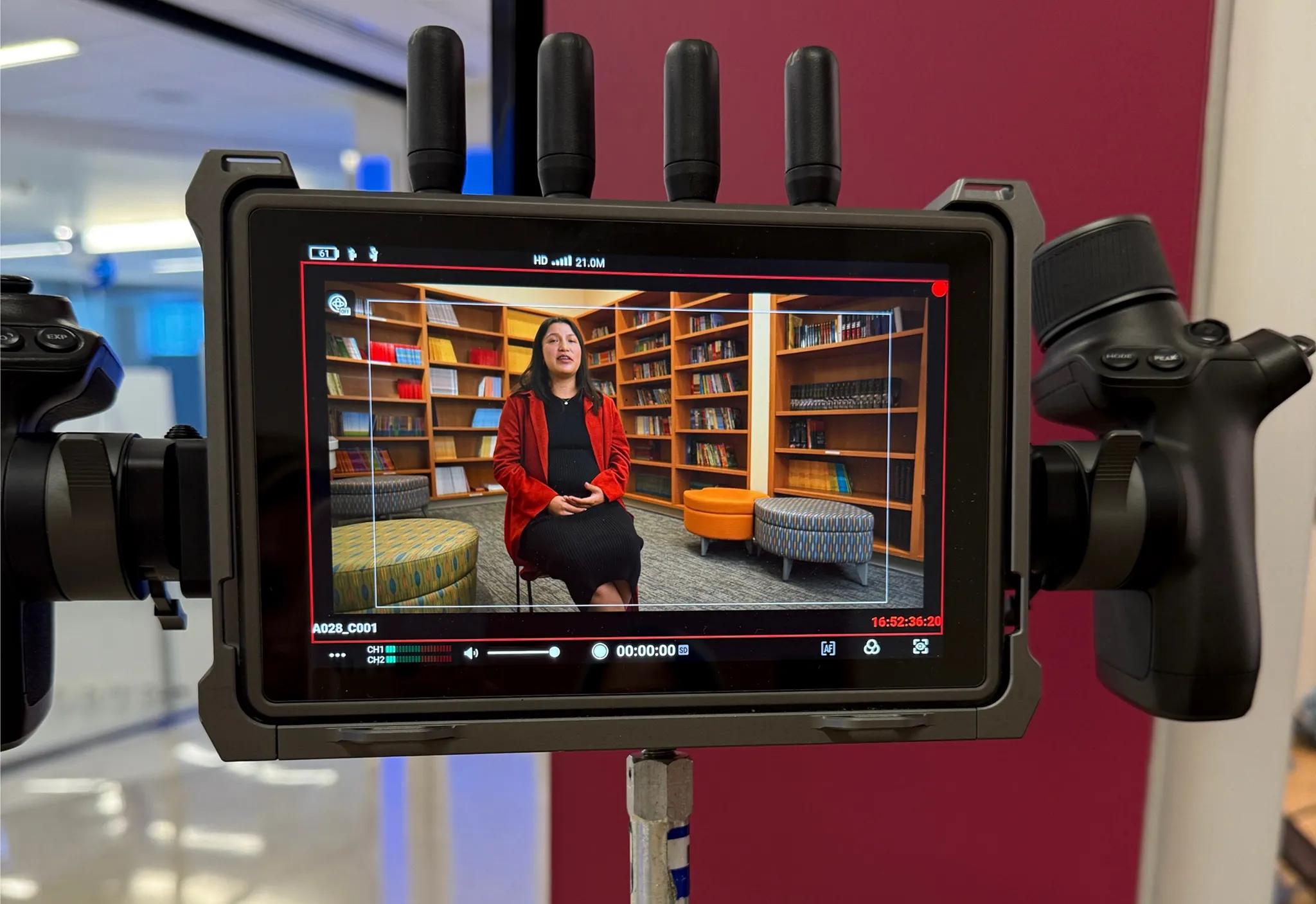 Camera monitor displays a woman in a red jacket being filmed seated in a library setting.