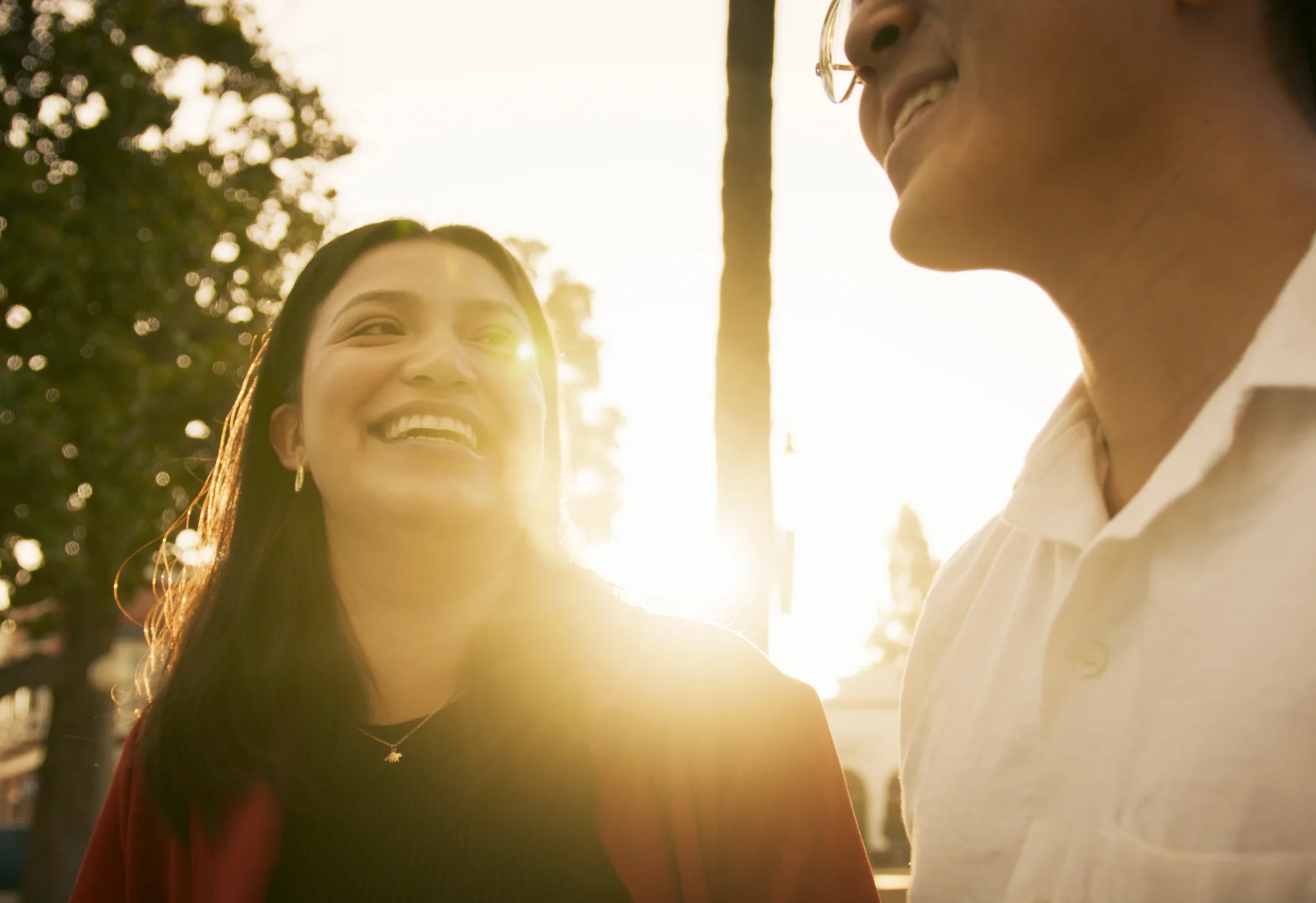 Young woman laughs with a friend in glasses at golden hour, warm sun flaring behind them.