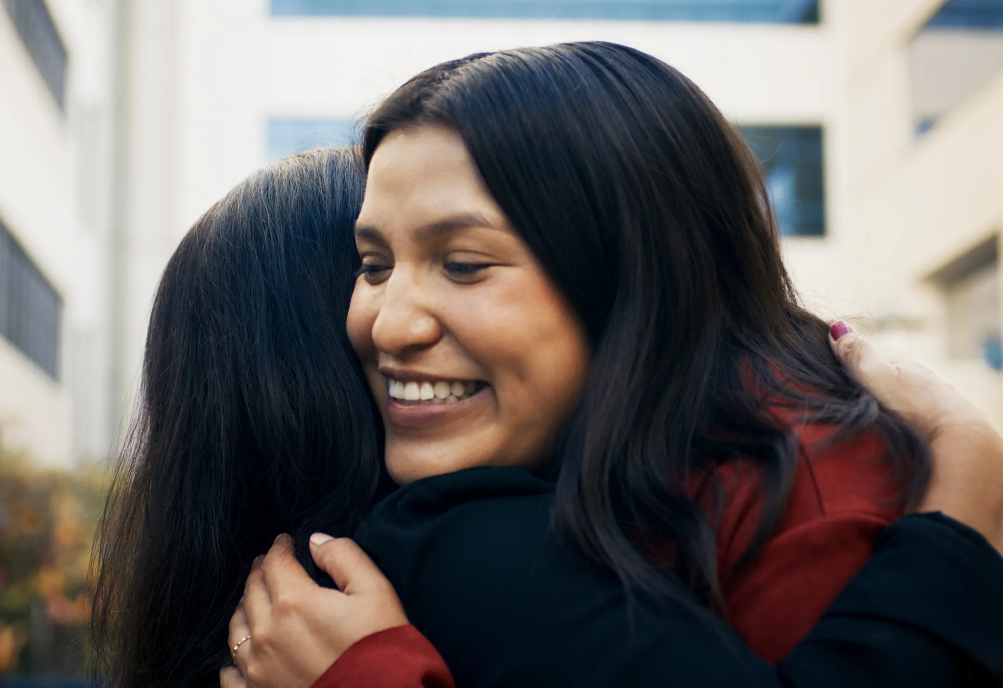 Two women share a warm embrace outdoors, one smiling brightly over the other's shoulder.