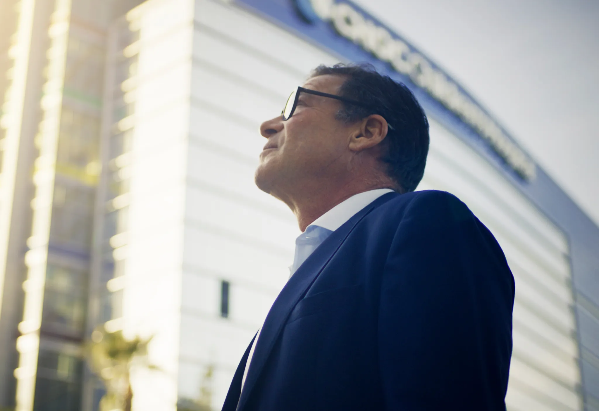 Man in dark blue suit and glasses looks upward toward a sunlit hospital building facade.