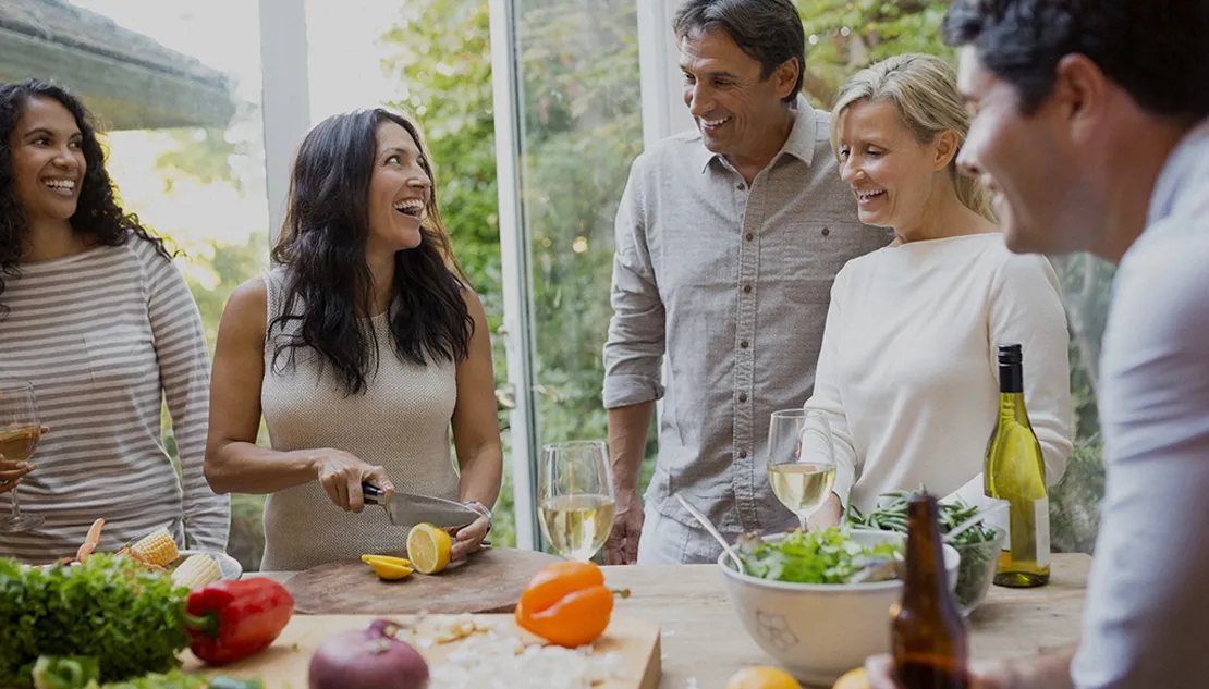 Group of friends laughing together while preparing a meal with fresh vegetables, wine, and salad in a Shea Homes kitchen.