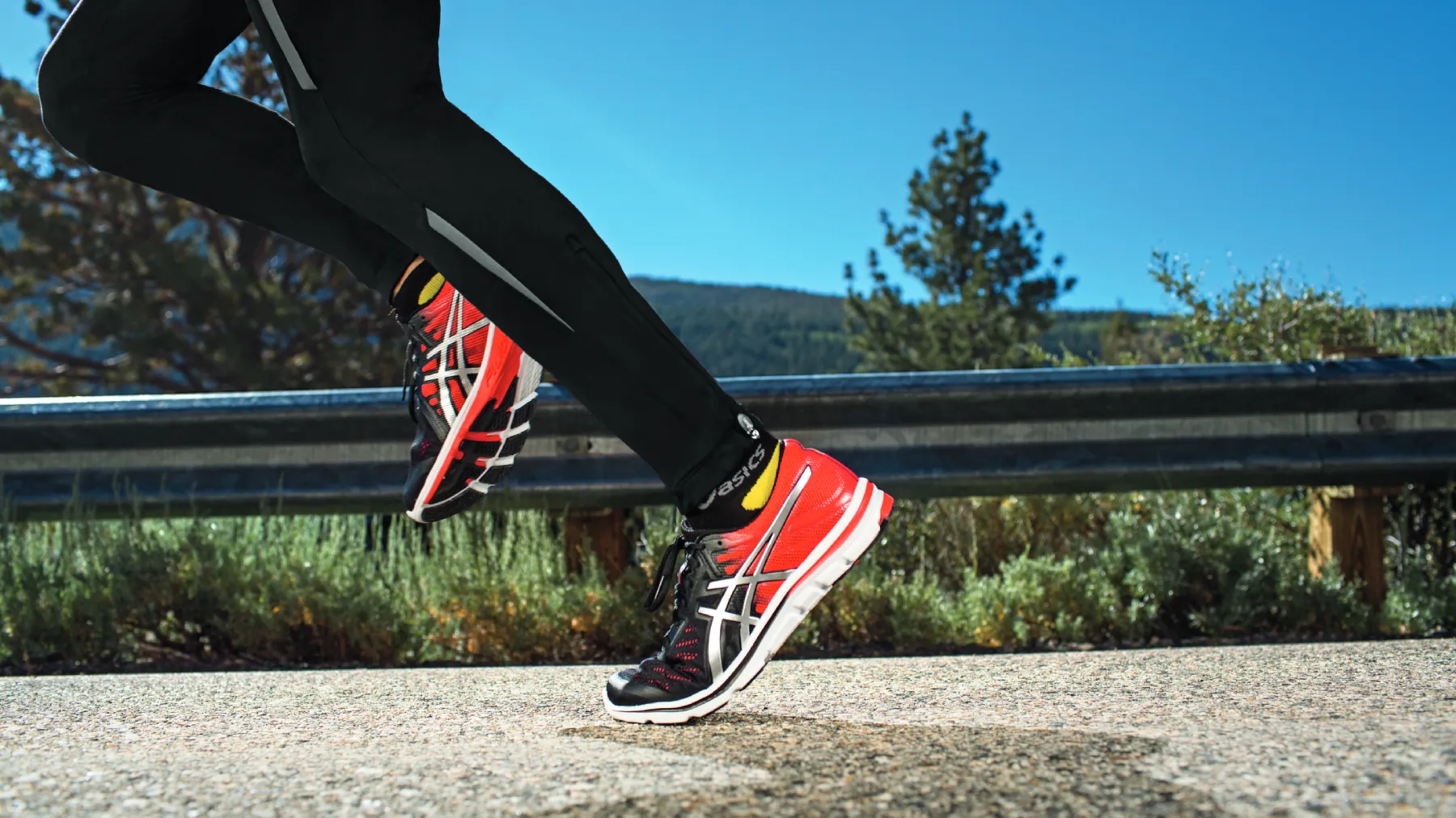 Runner's feet in red and black ASICS shoes mid-stride on a road with mountains and blue sky in the background