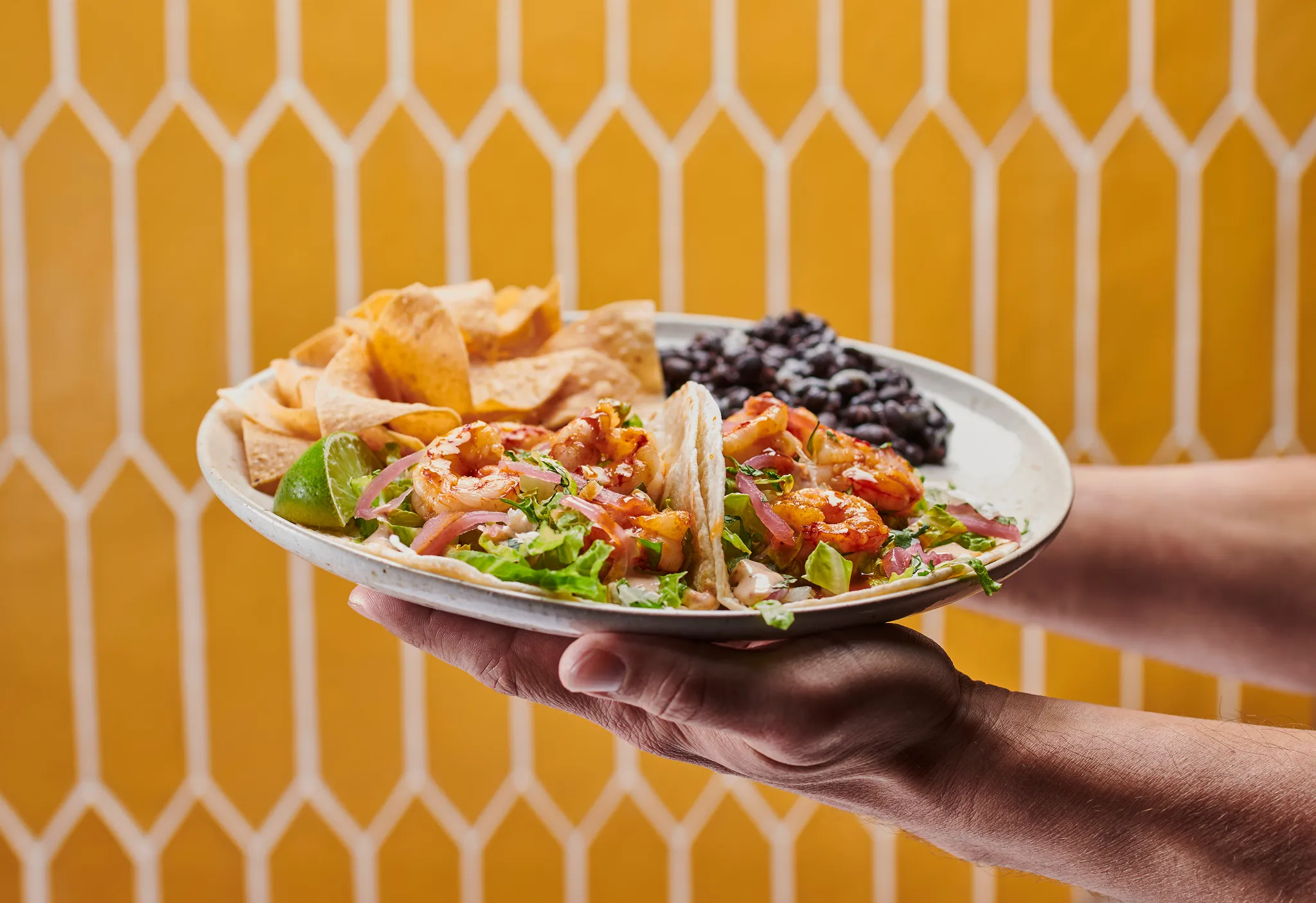 Hand holding a plate of Rubio's shrimp tacos with chips and black beans in front of yellow tile wall.