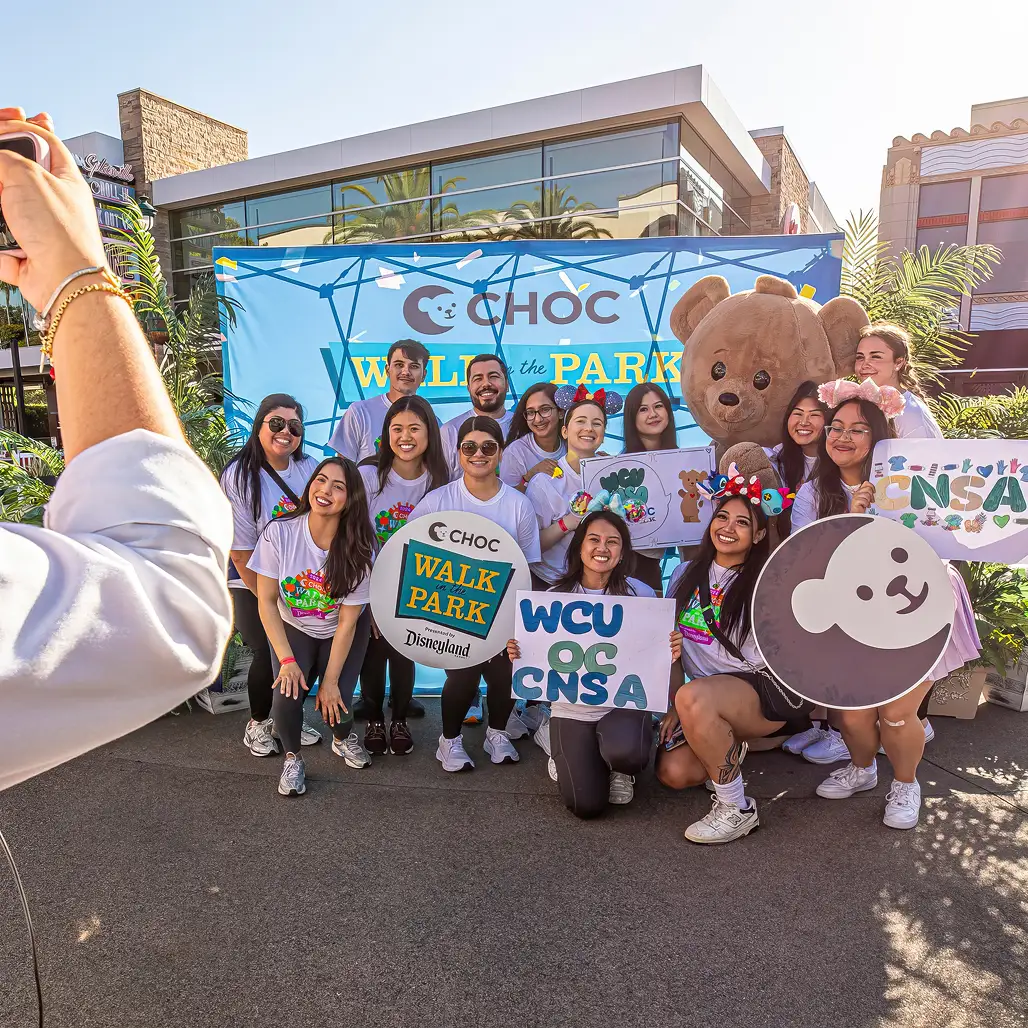  A photographer snaps student volunteers in CHOC Walk shirts holding team signs, posed with the CHOC bear mascot in front of the event's branded backdrop. 