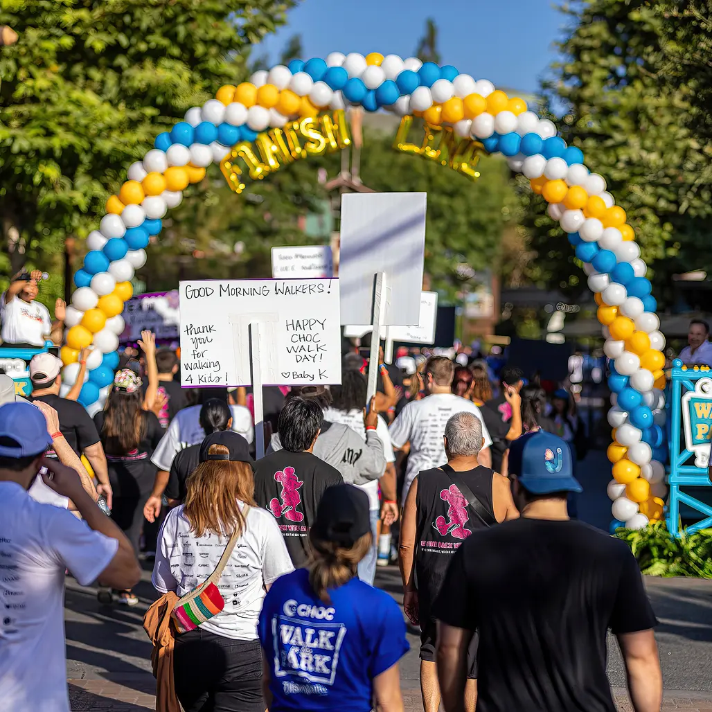 Walkers stream toward a blue, yellow, and white balloon finish-line arch at CHOC Walk, some carrying handmade "Good Morning Walkers!" and "Happy CHOC Walk Day!" signs.