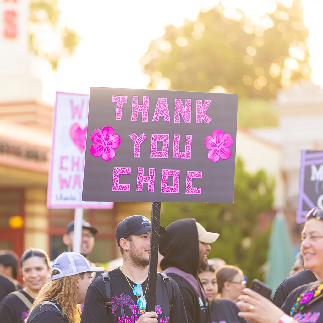 Close-up of a participant holding a handmade "Thank You CHOC" sign decorated with pink hibiscus flowers, surrounded by smiling walkers in matching team tees at golden hour.