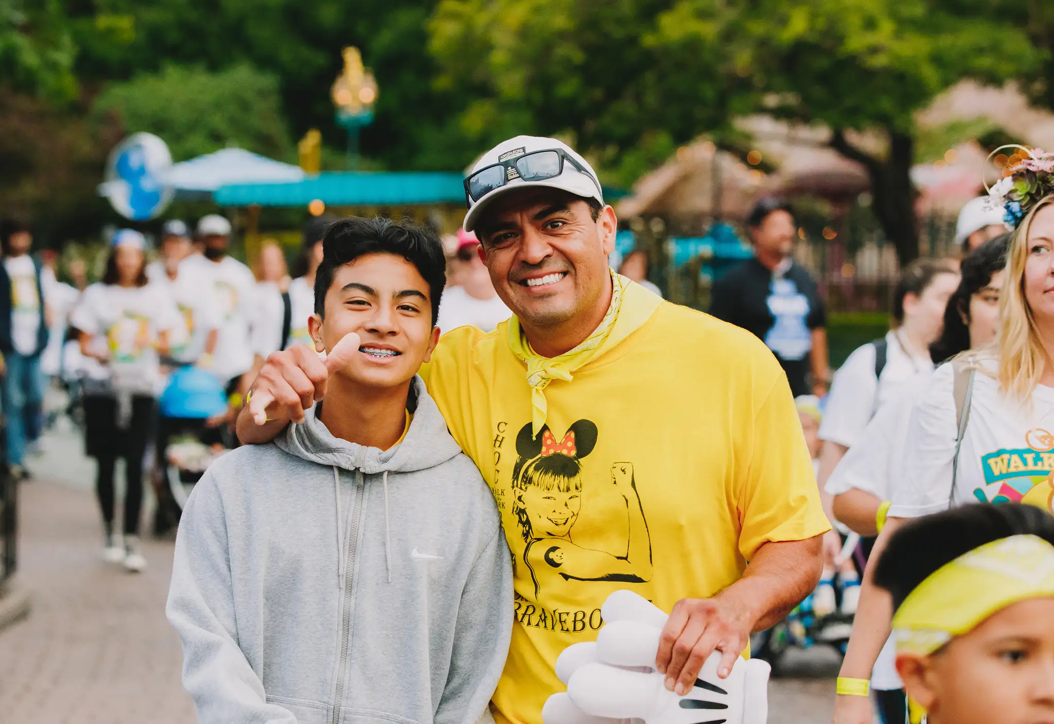 A smiling father and teenage son pose together at the CHOC Walk in the Park, the dad wearing a yellow event t-shirt as crowds of walkers move past behind them. 