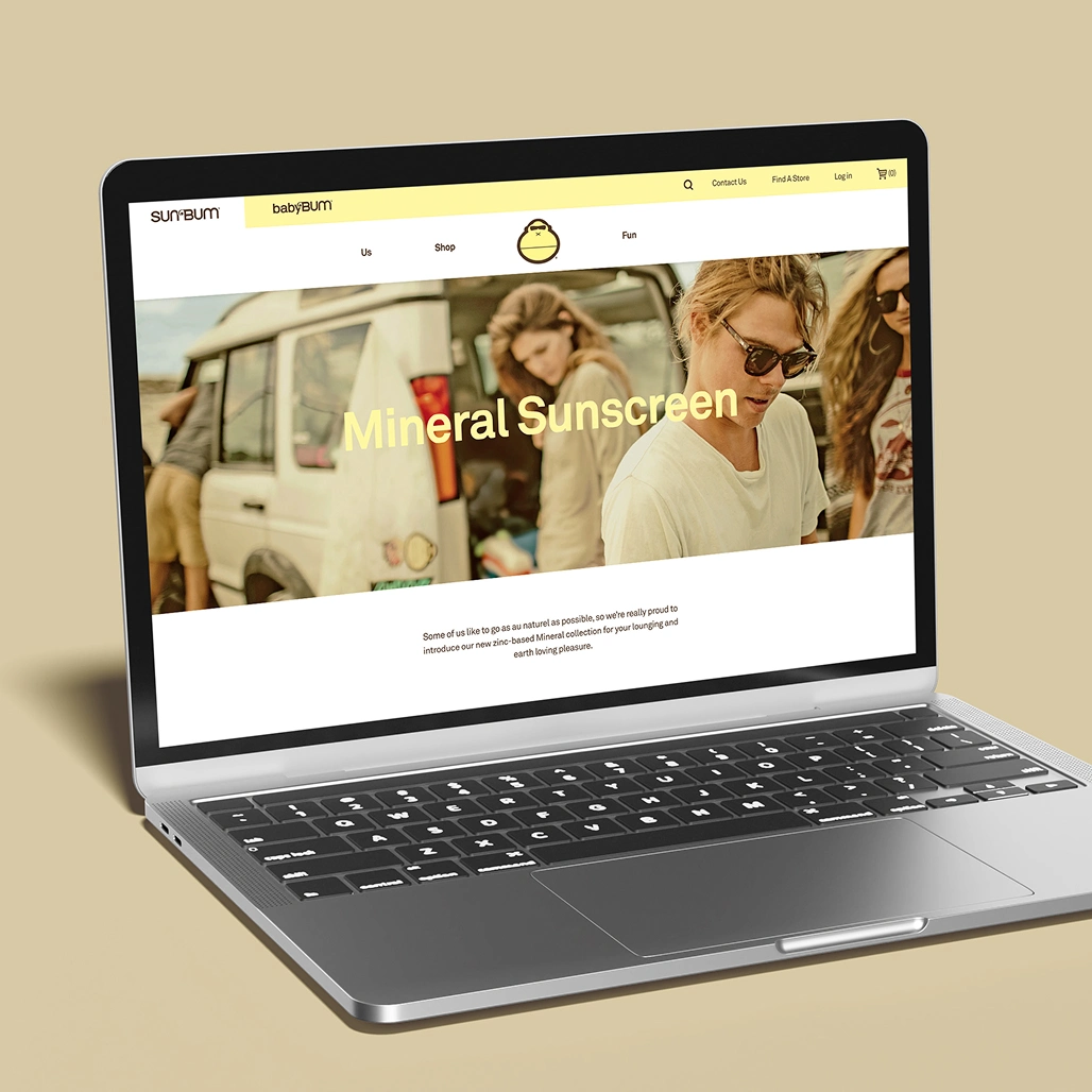 A laptop displaying the Sun Bum website's Mineral Sunscreen landing page, featuring a hero image of beachgoers near a van and introductory product copy.