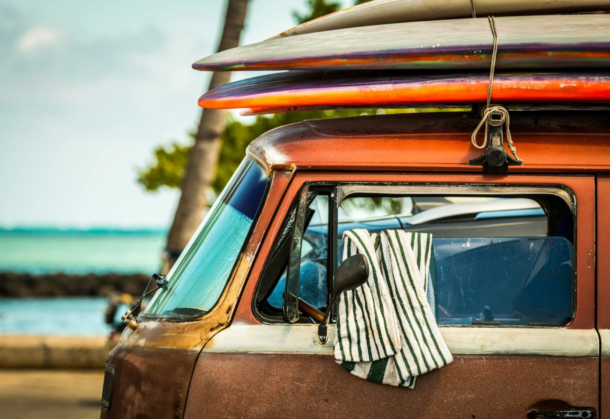 Close-up of the cab of a weathered vintage van with colorful surfboards strapped to the roof, parked near a tropical beach with palm trees in the background.