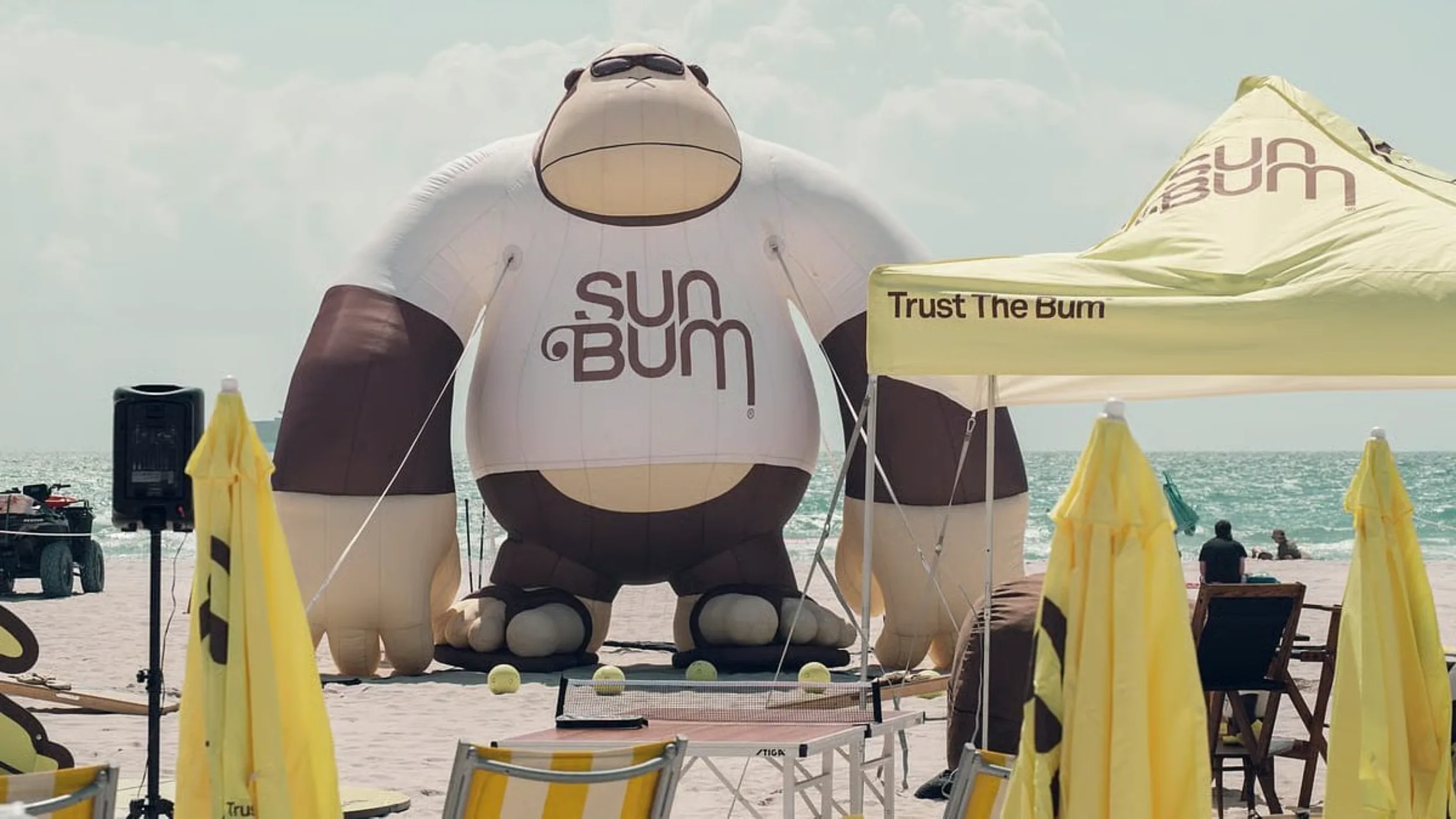 Giant inflatable Sun Bum gorilla mascot towering over a beach setup with umbrellas and a "Trust the Bum" tent