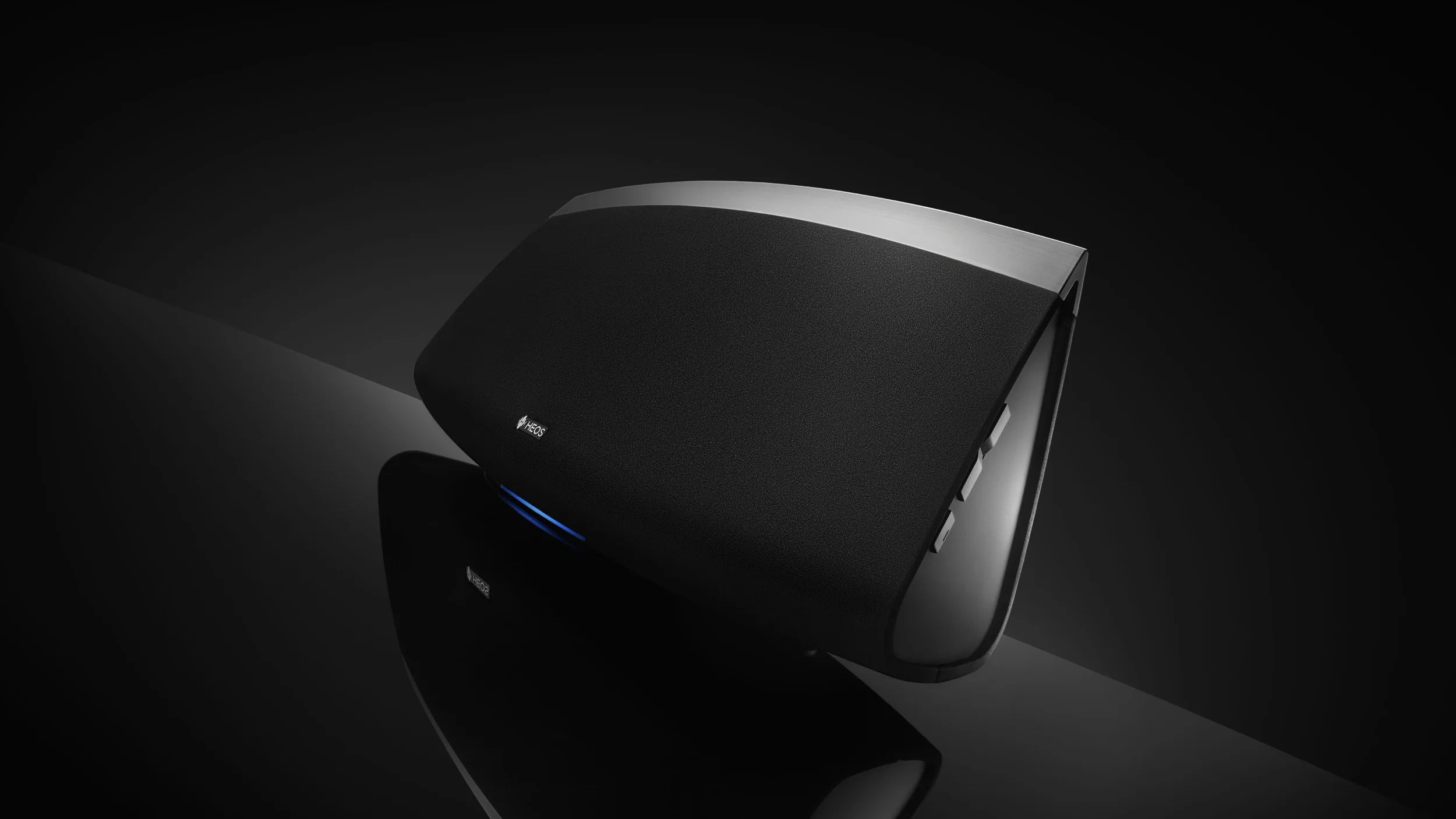 HEOS by Denon wireless speaker in black with brushed metal trim and blue glowing light on a dark reflective surface