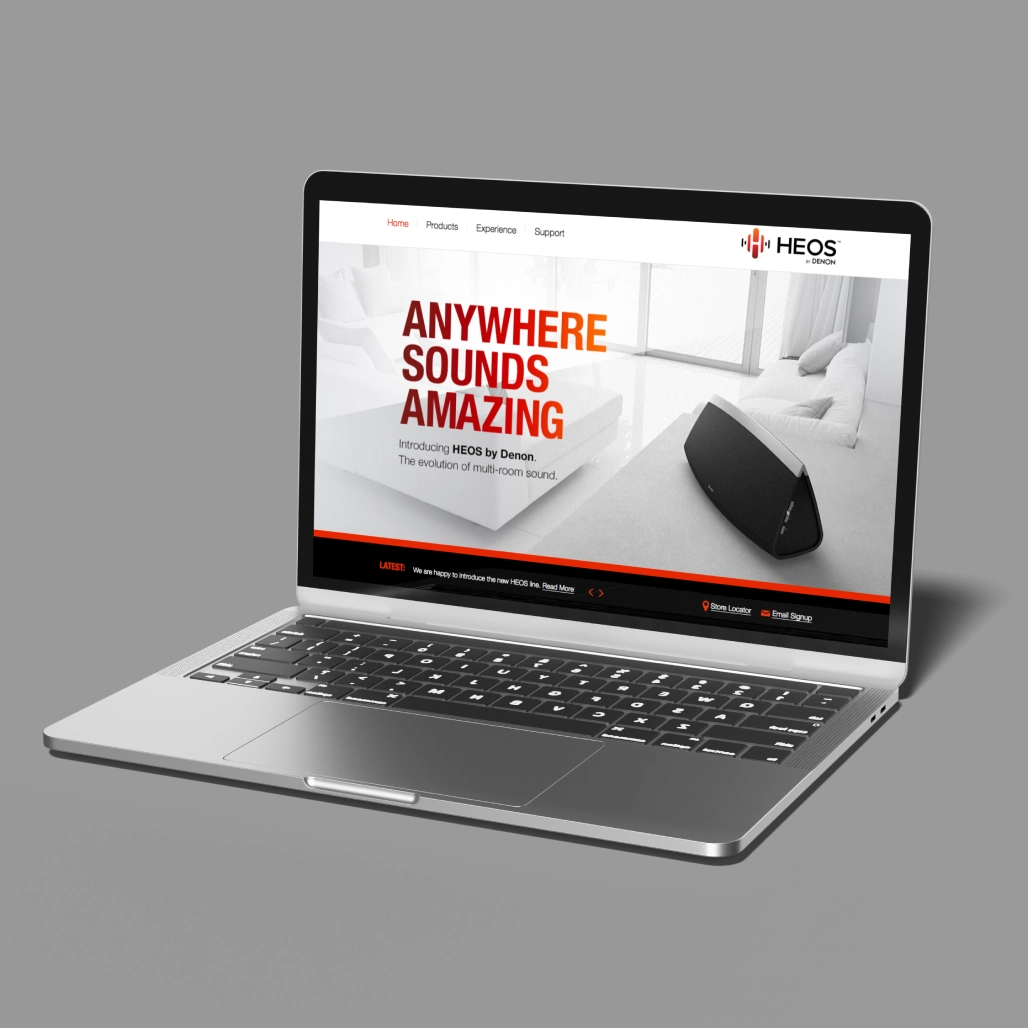 Laptop displaying HEOS by Denon homepage with "Anywhere Sounds Amazing" headline and speaker in a bright living room
