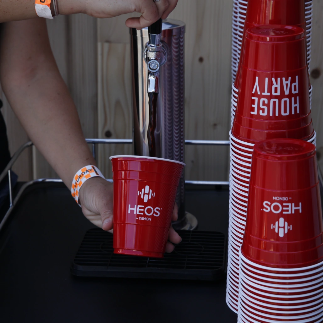 Hand pouring a drink into a red HEOS by Denon cup beside stacked House Party cups at a festival tap