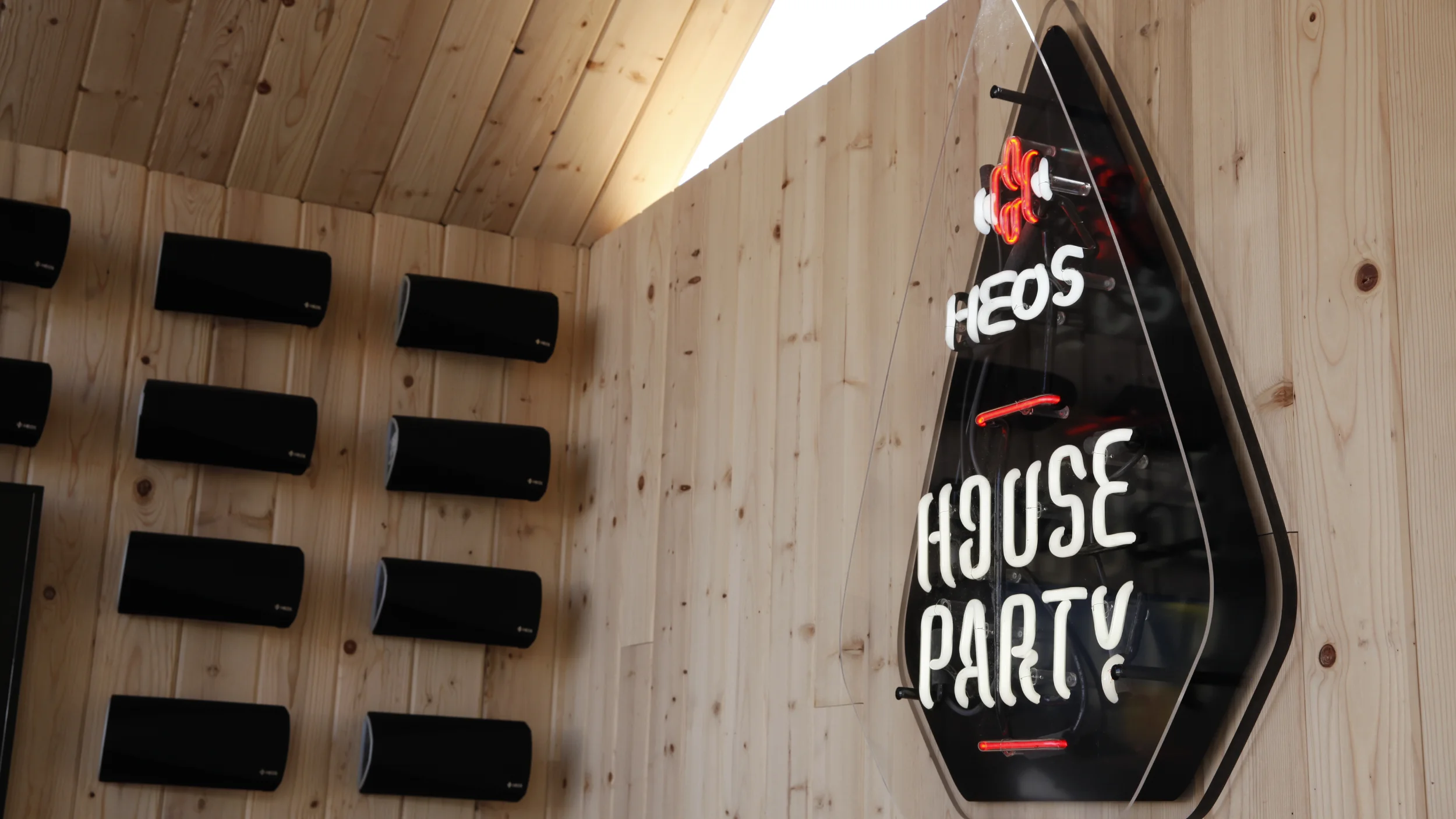 Neon HEOS House Party sign beside rows of black HEOS speakers mounted on a light wood wall
