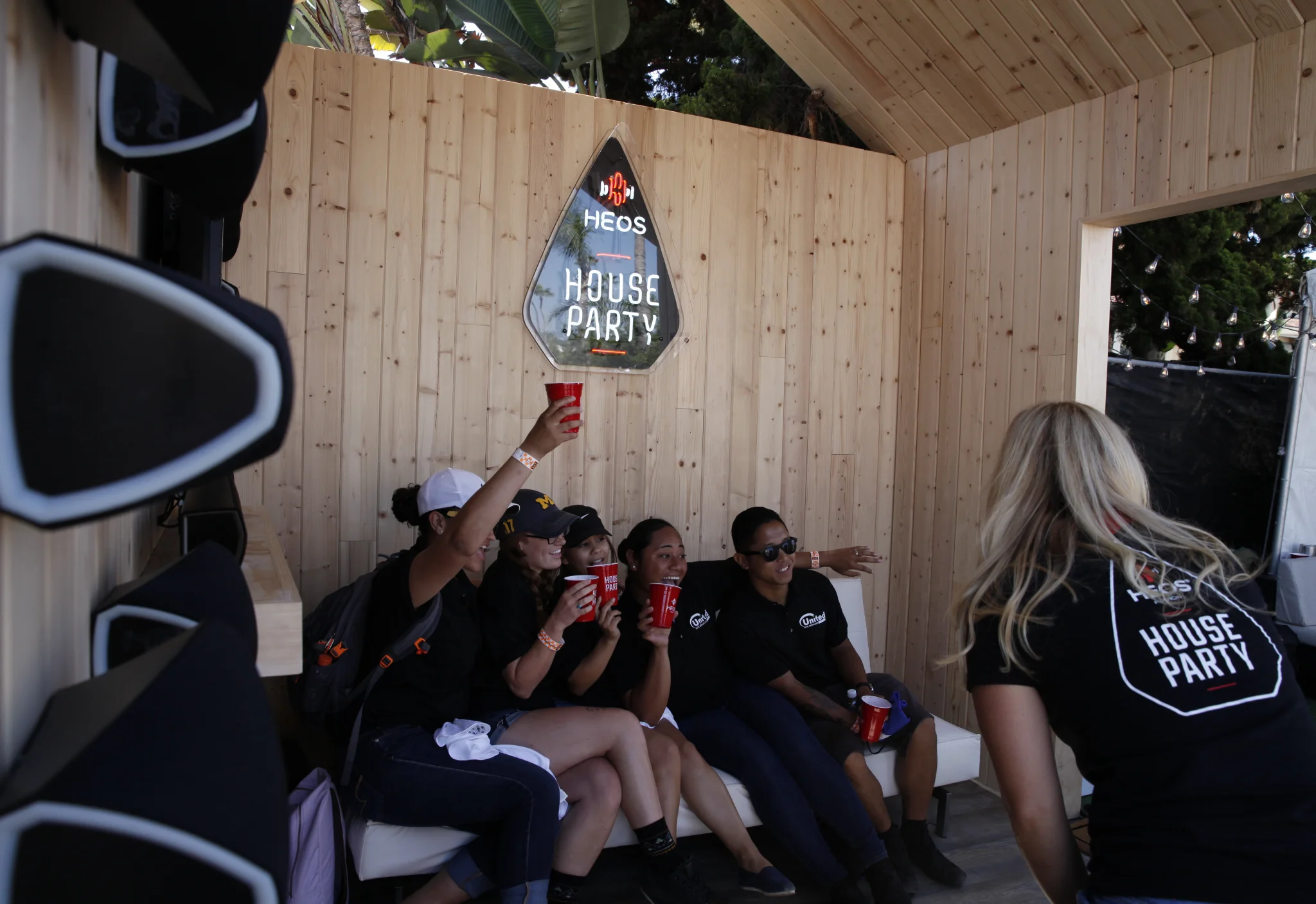 Group posing with red cups on a white couch for a photo booth shot in the wood-paneled HEOS House Party lounge
