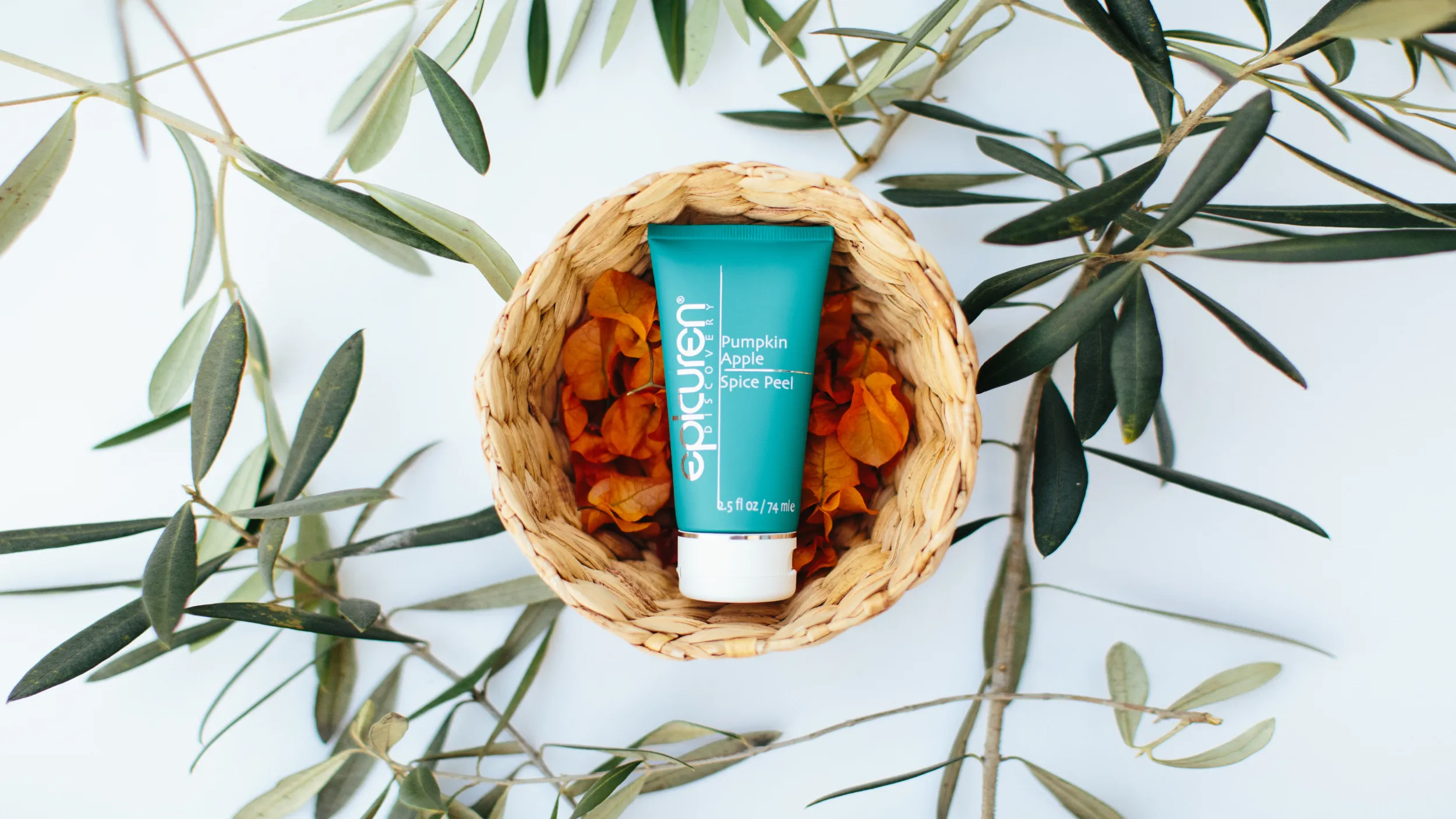 Overhead view of Epicuren Pumpkin Apple Spice Peel in a woven basket with orange petals and olive branches on white