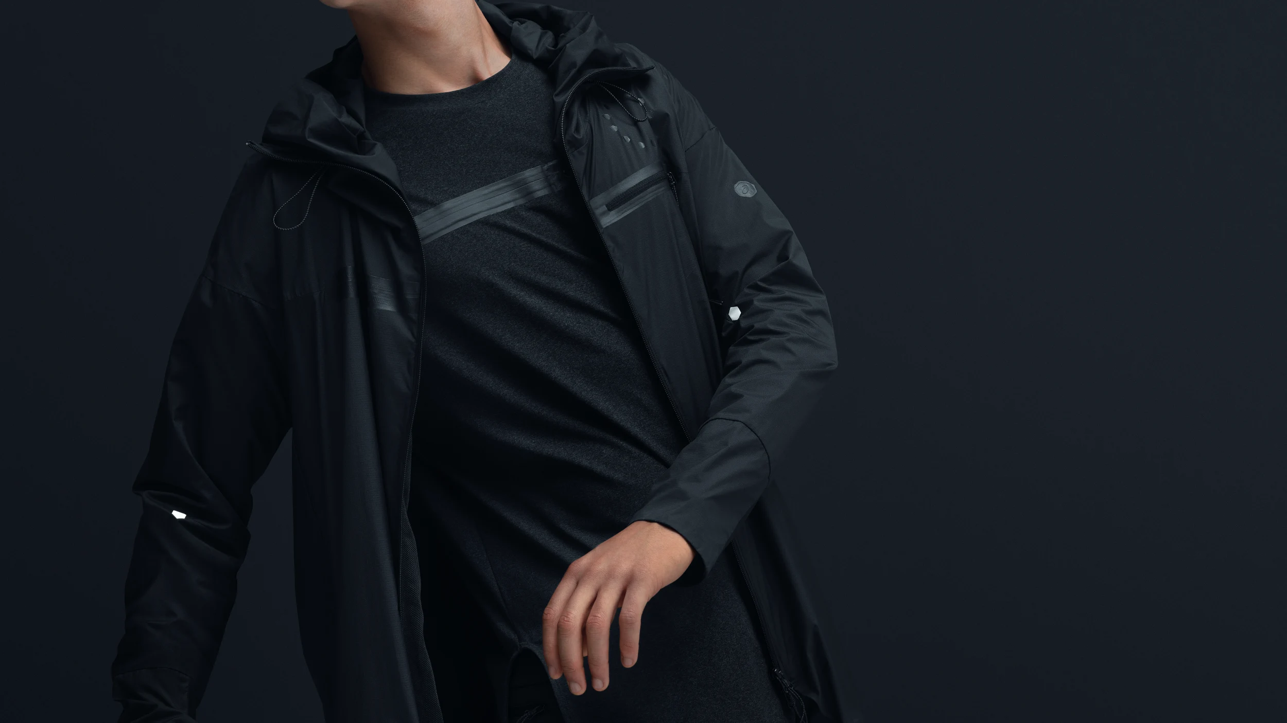 Model in a black ASICS JYUNI Black Collection hooded jacket over a dark heathered shirt against a dark backdrop