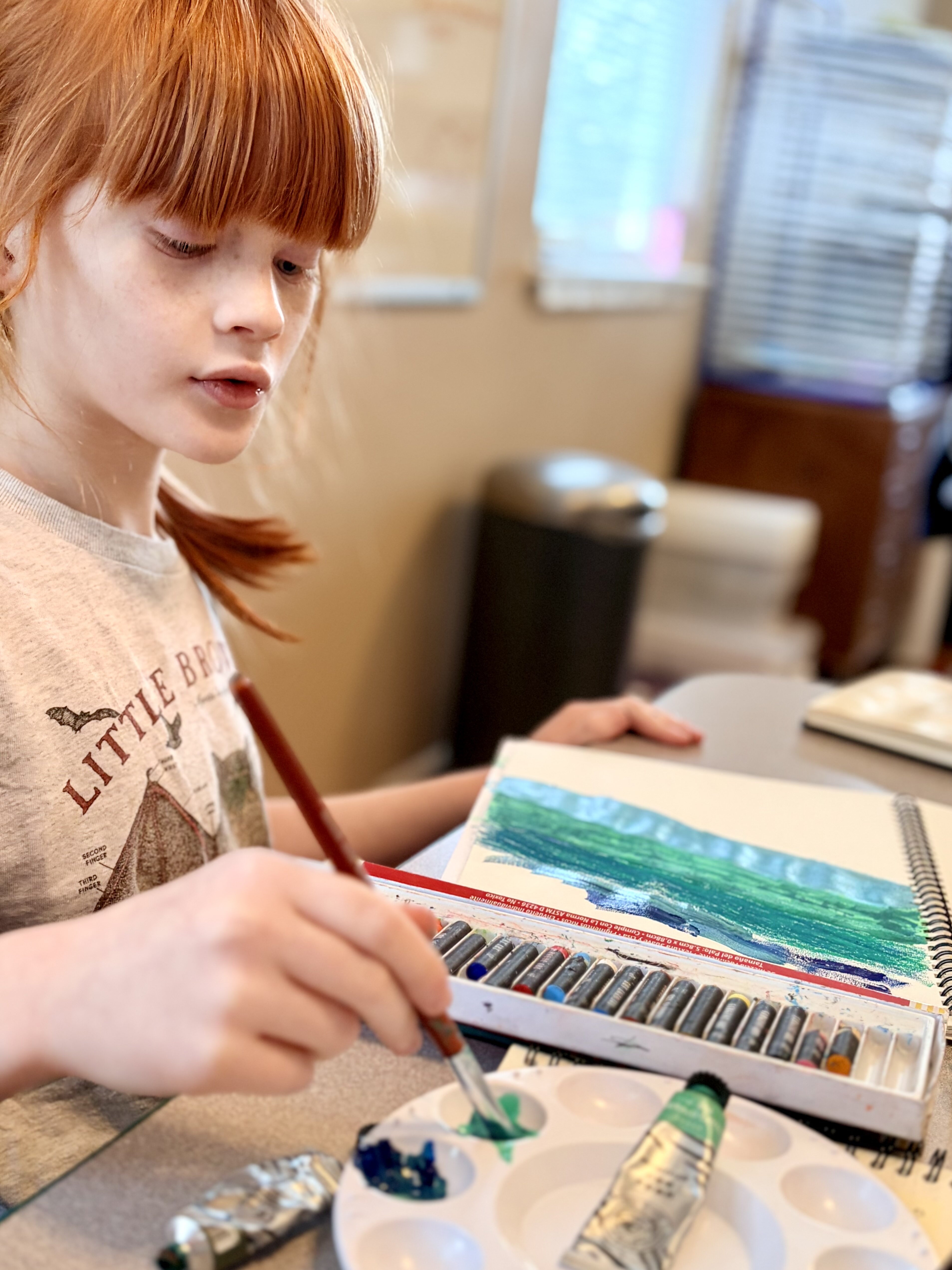 Young red-haired girl painting with blue and green colors on a sketchbook at a table.