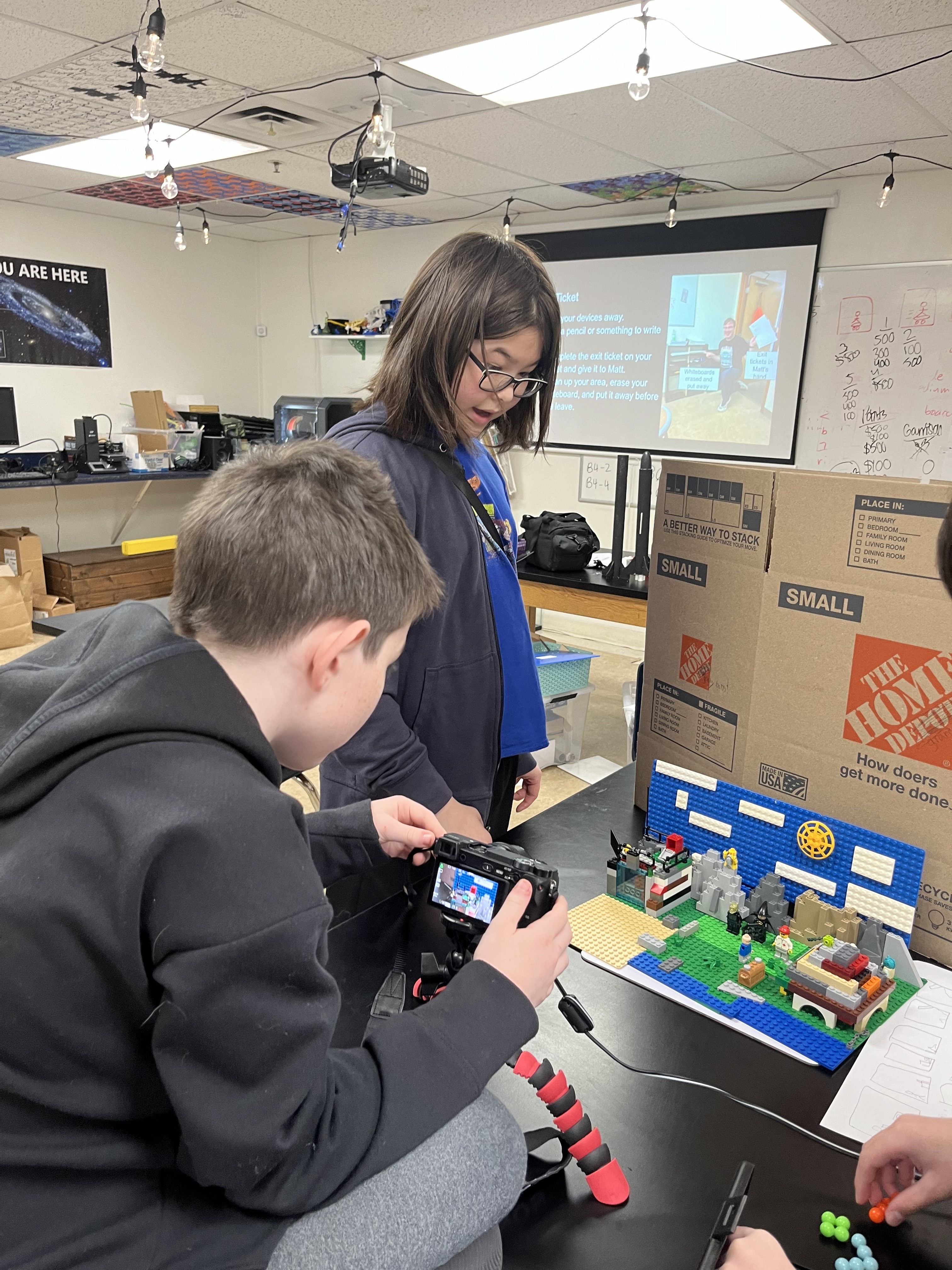 Two children in a classroom using a camera to photograph a LEGO setup on a table.