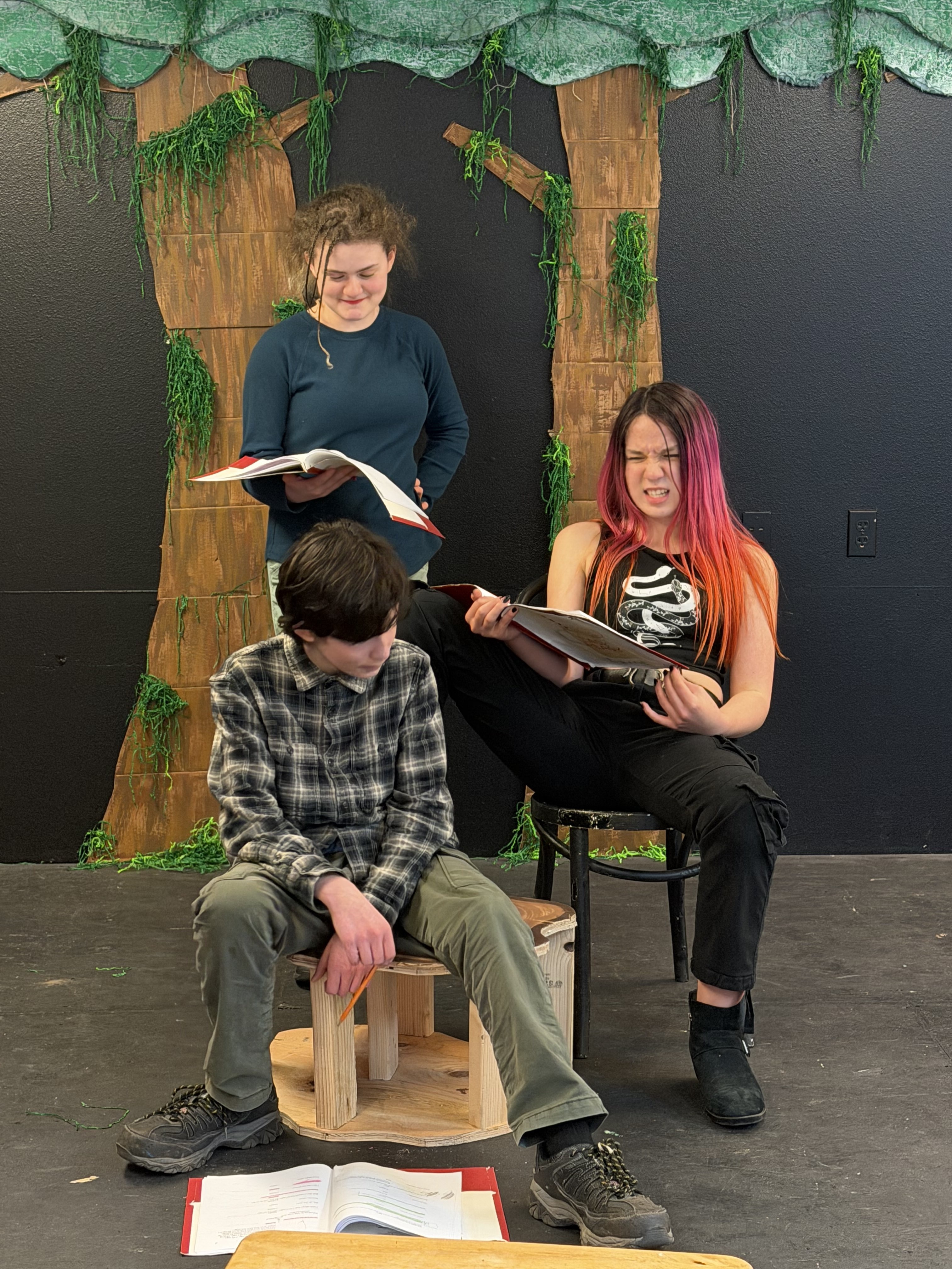 Three teens sitting and standing indoors with books, one girl reading and making a face, boy and another girl holding open books, against a black wall decorated with tree and greenery artwork.