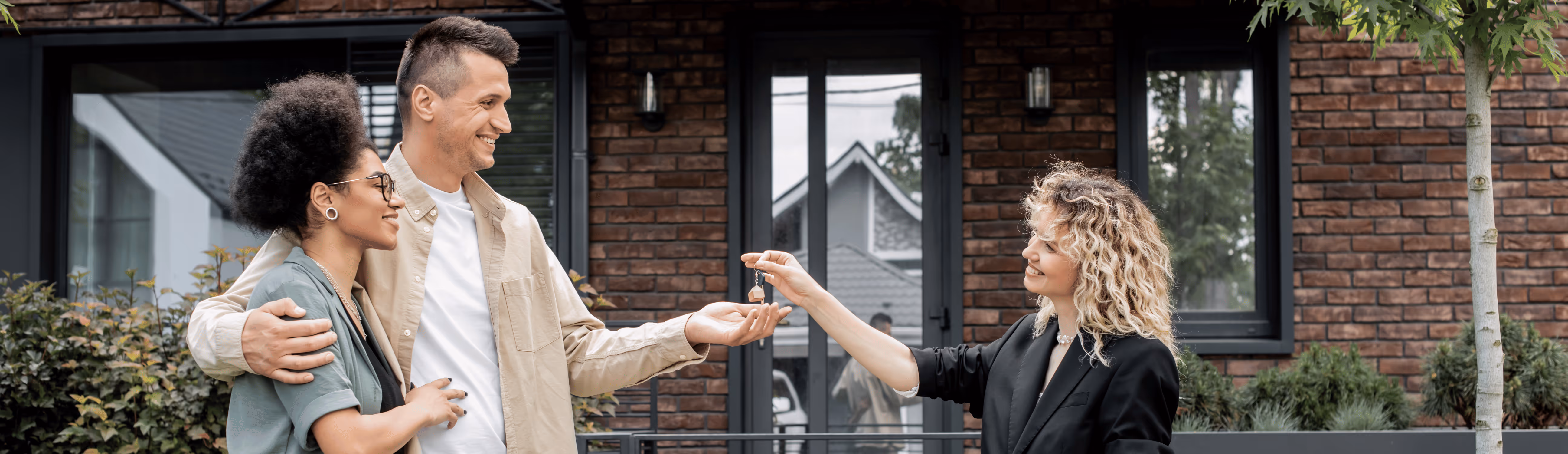 Smiling couple receiving house keys from a real estate agent outside a modern brick house.