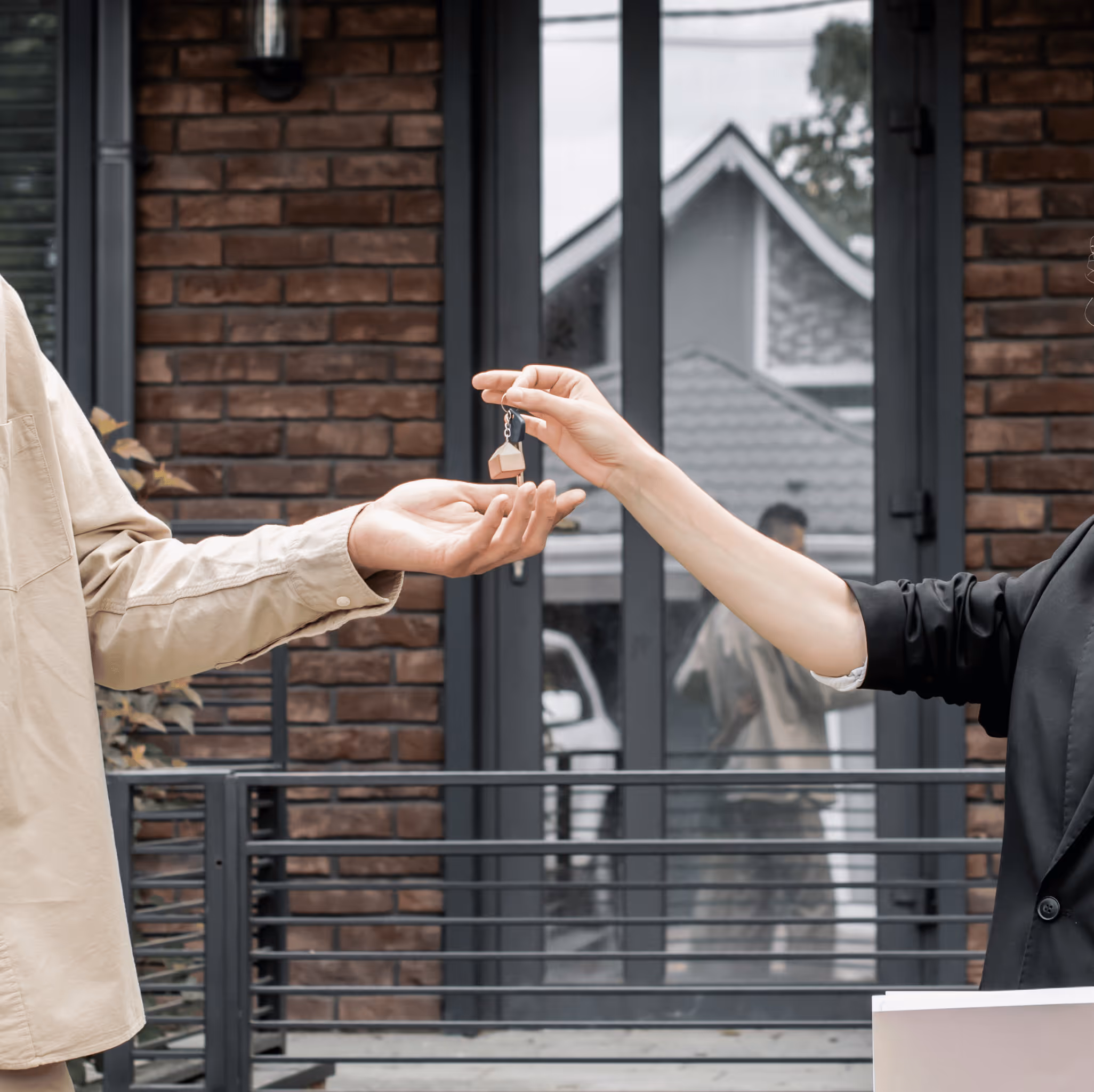 Person handing over a house-shaped keychain to another person in front of a brick building with windows.
