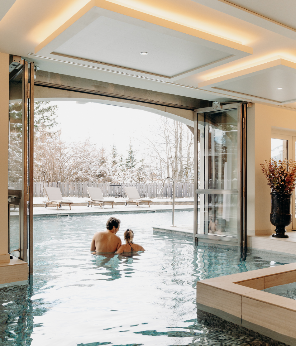 Couple dans la piscine intérieure et extérieure du spa face au paysage enneigé aux Chalets du Mont d’Arbois — Megève.