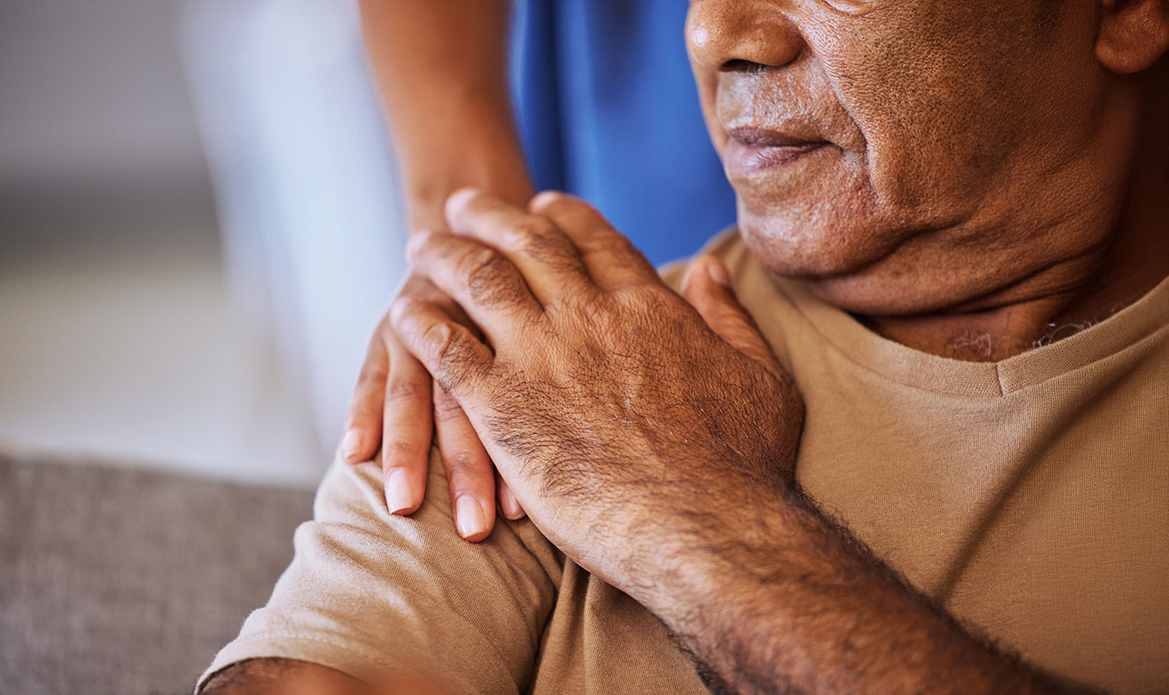 A seated woman, only partially visible in the frame. Another person behind her has their hand on her shoulder; the seated woman's hand has met the other's hand. Suggests caring, support, and/or help.