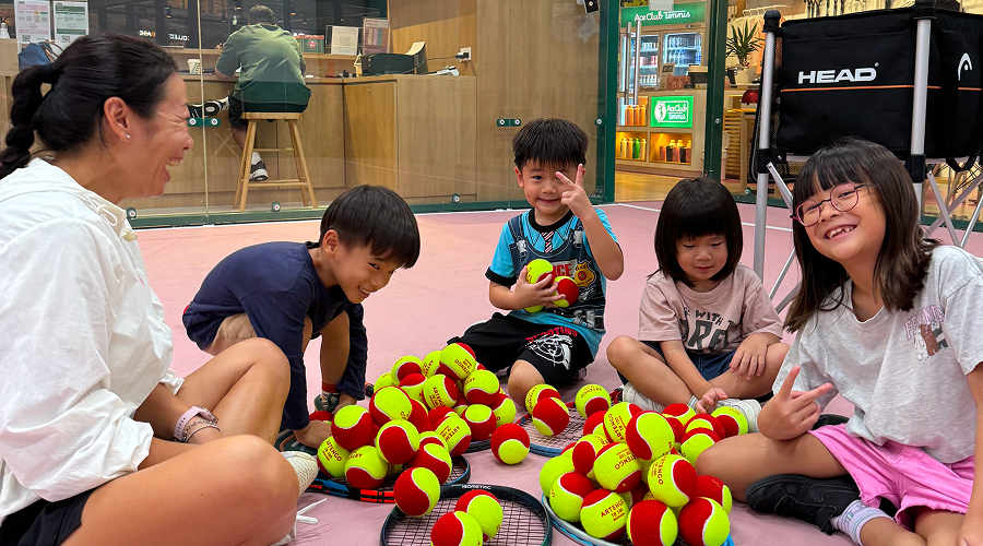 Kids with a bunch of tennis balls and racquets in an indoor court.