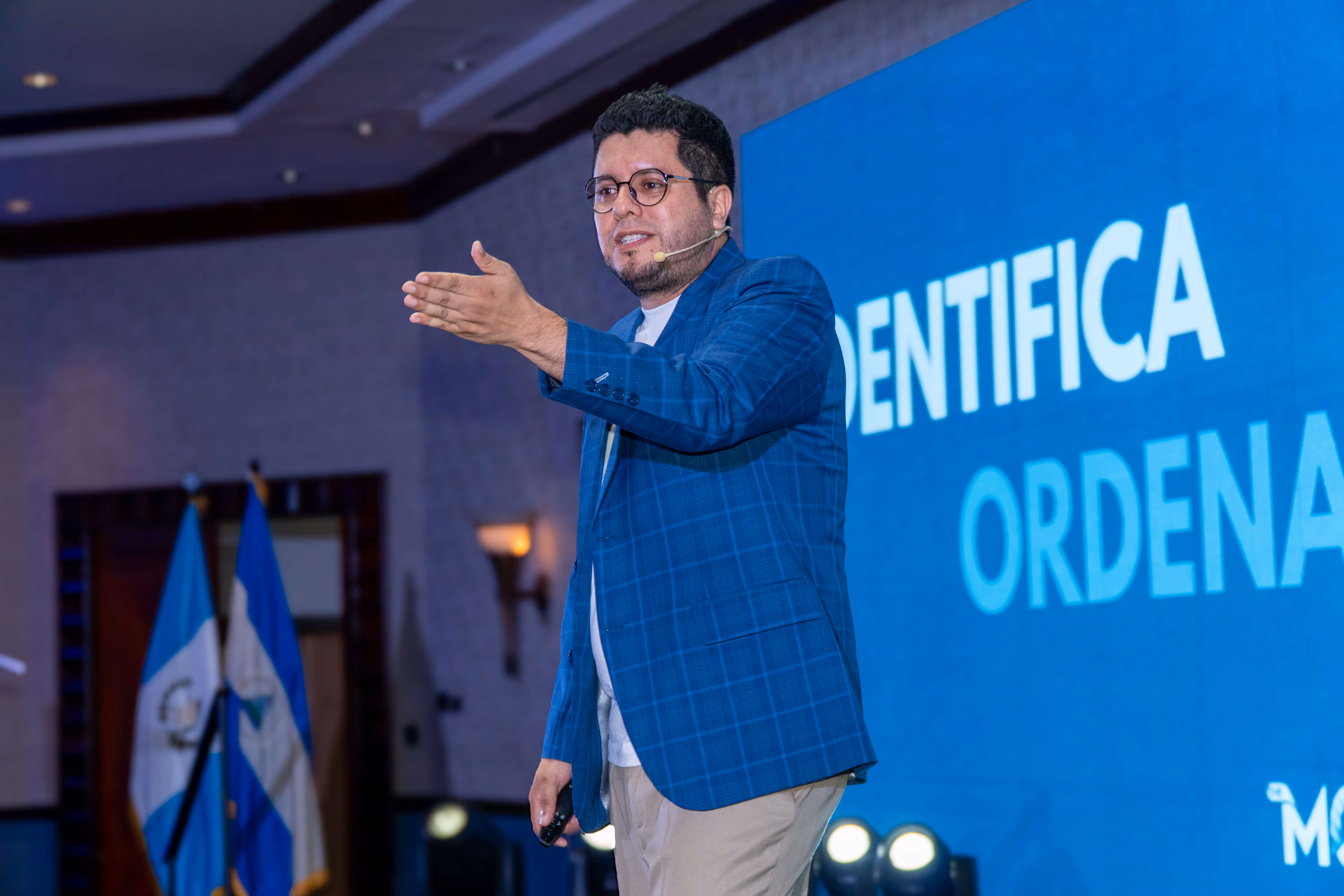 Hombre con micrófono de diadema y chaqueta azul hablando en un escenario con dos banderas de Guatemala al fondo.