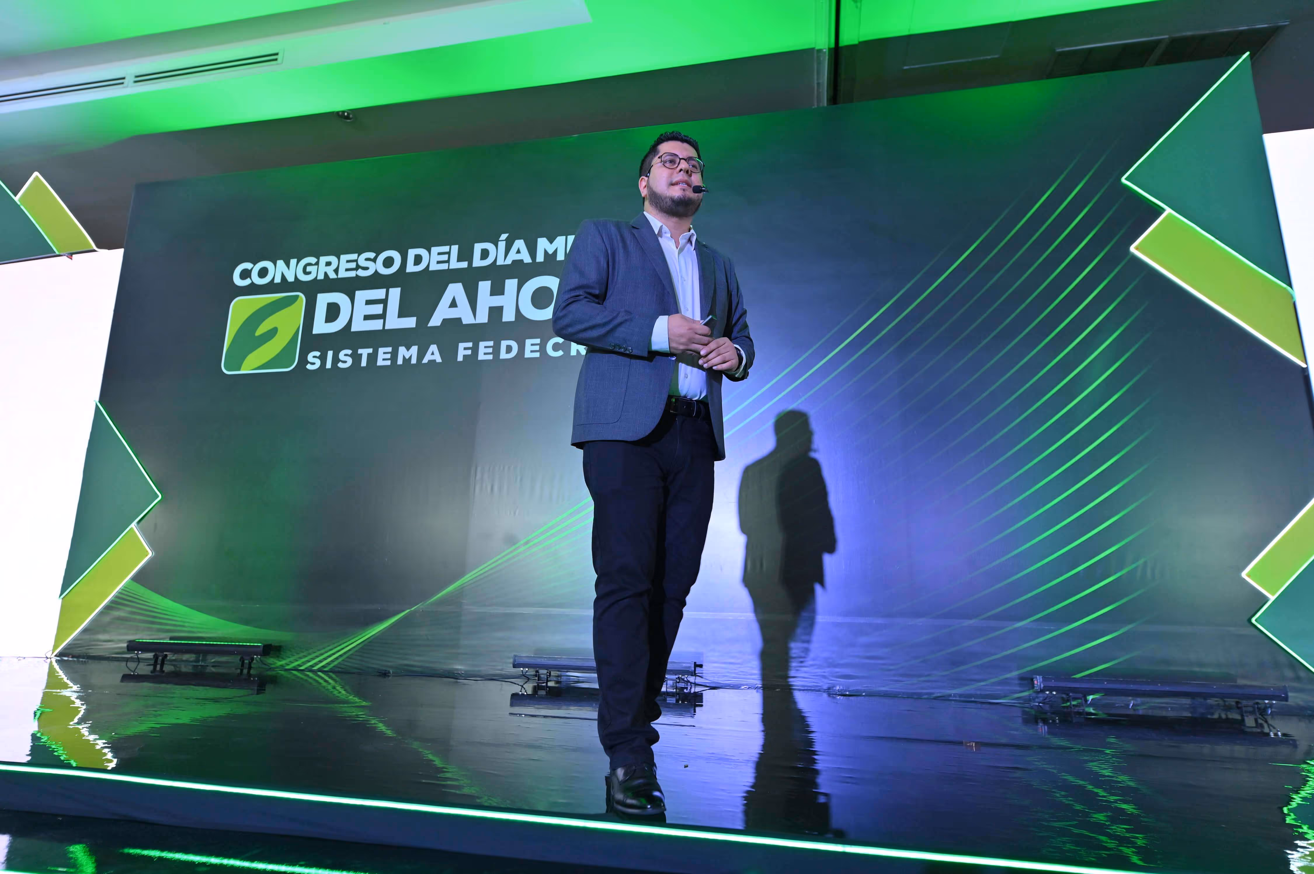 Hombre con chaqueta y micrófono de diadema hablando en un escenario con fondo negro y verde del Congreso del Día Mundial del Ahorro.