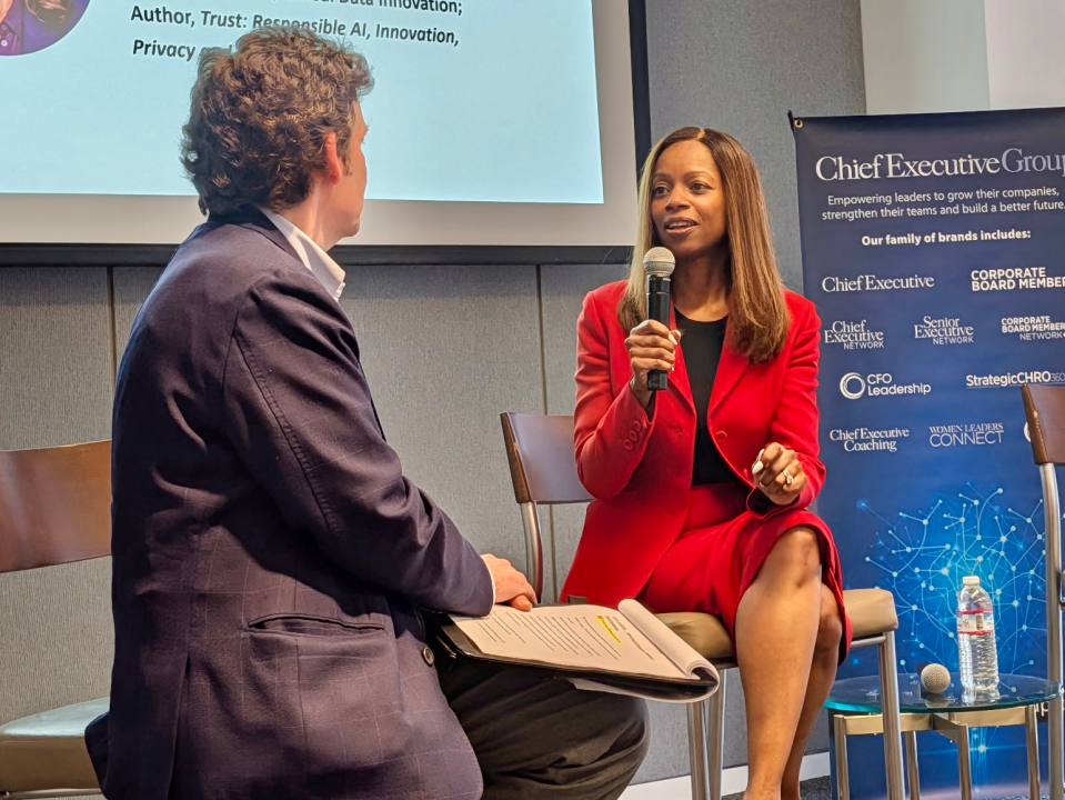 Woman in a red suit holding a microphone speaking during a discussion with a man in a dark blazer holding notes.