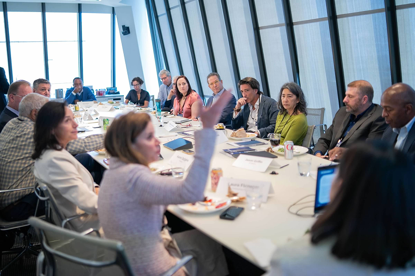 Group of professionals engaged in a meeting around a large conference table in a modern office with tall windows.
