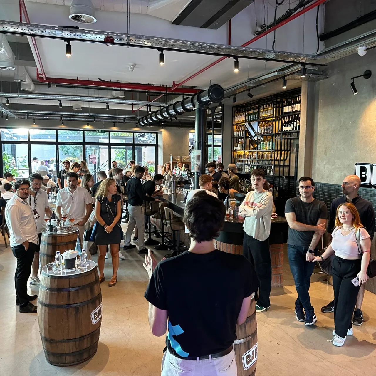 A presentation help by a guys with curly hair with a black T-shirt and on his left side showing the X logo of MultiversX. The crowd is in a modern networking spot with barrels as tables and bar.