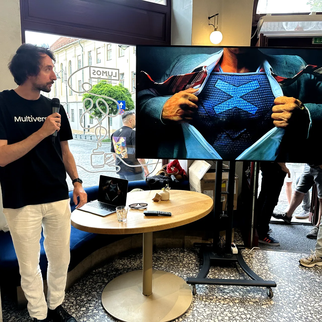 A curly haired person with MultiversX Logo on his t-shirt presenting a slide on a big monitor depicting a Superman like person showing his chest with a big blue X on it.