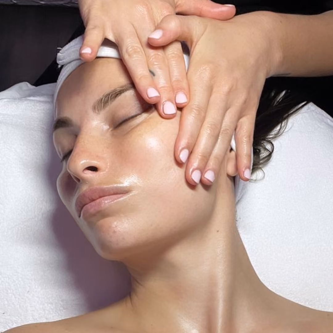 A woman with eyes closed receiving a facial massage with hands on her forehead and cheek.