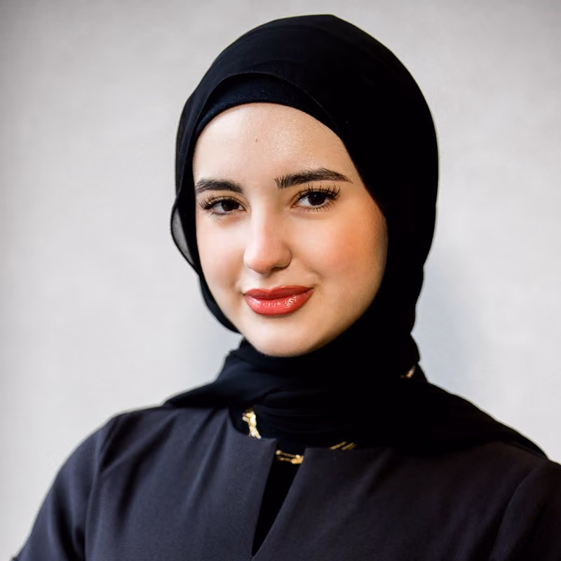 Portrait of a woman wearing a black hijab and black outfit, smiling softly against a neutral background.