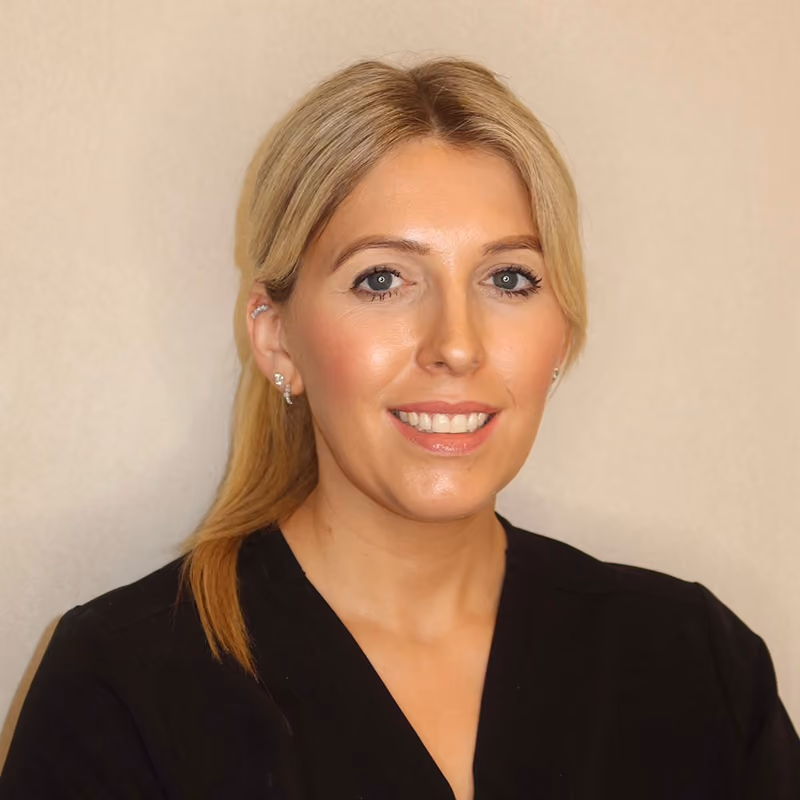 Smiling woman with blonde hair tied back wearing a black top against a beige background.